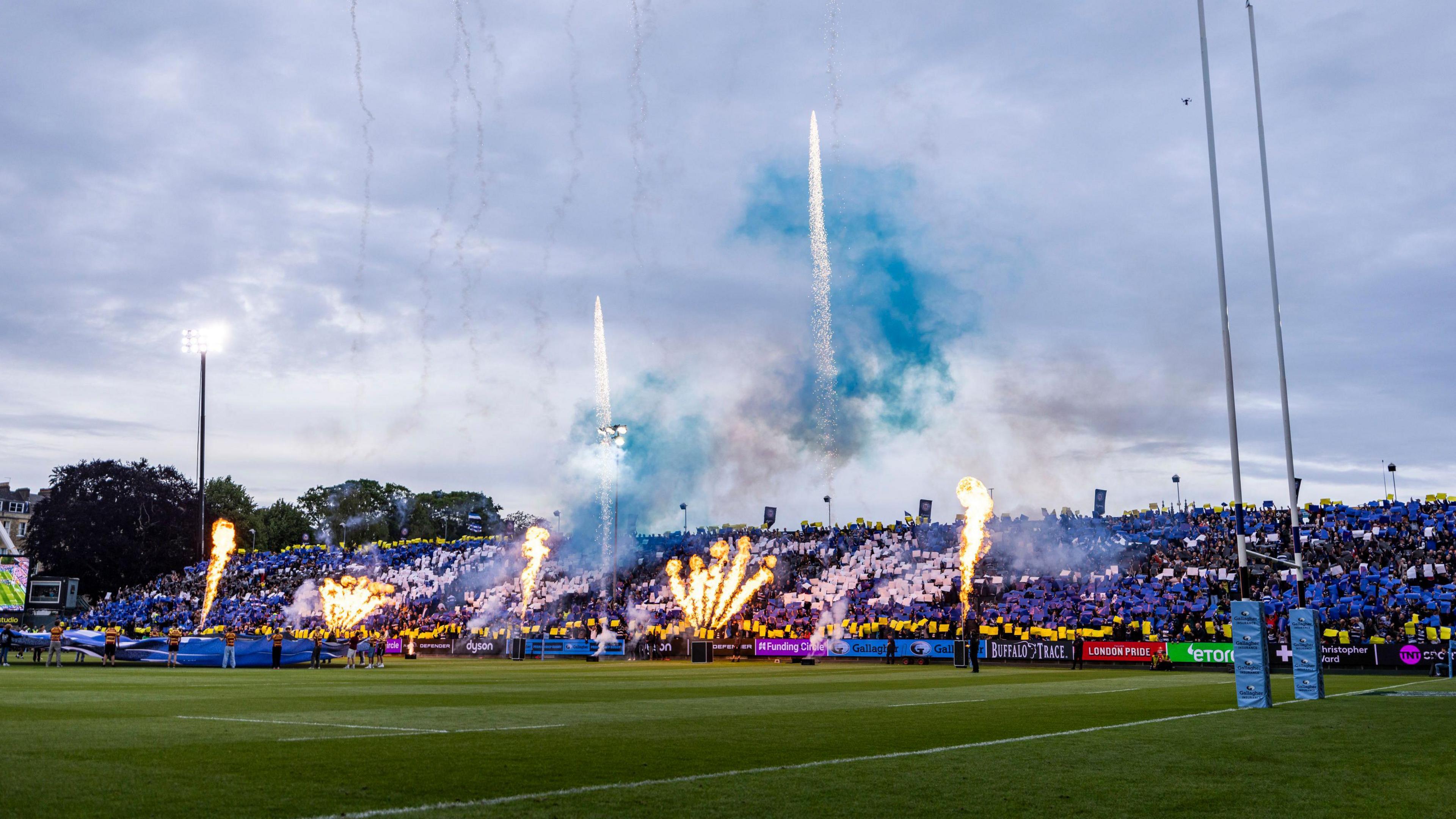 Fireworks explode on the side of the pitch and fans hold up coloured cards as the teams come out at The Rec for Bath v Bristol
