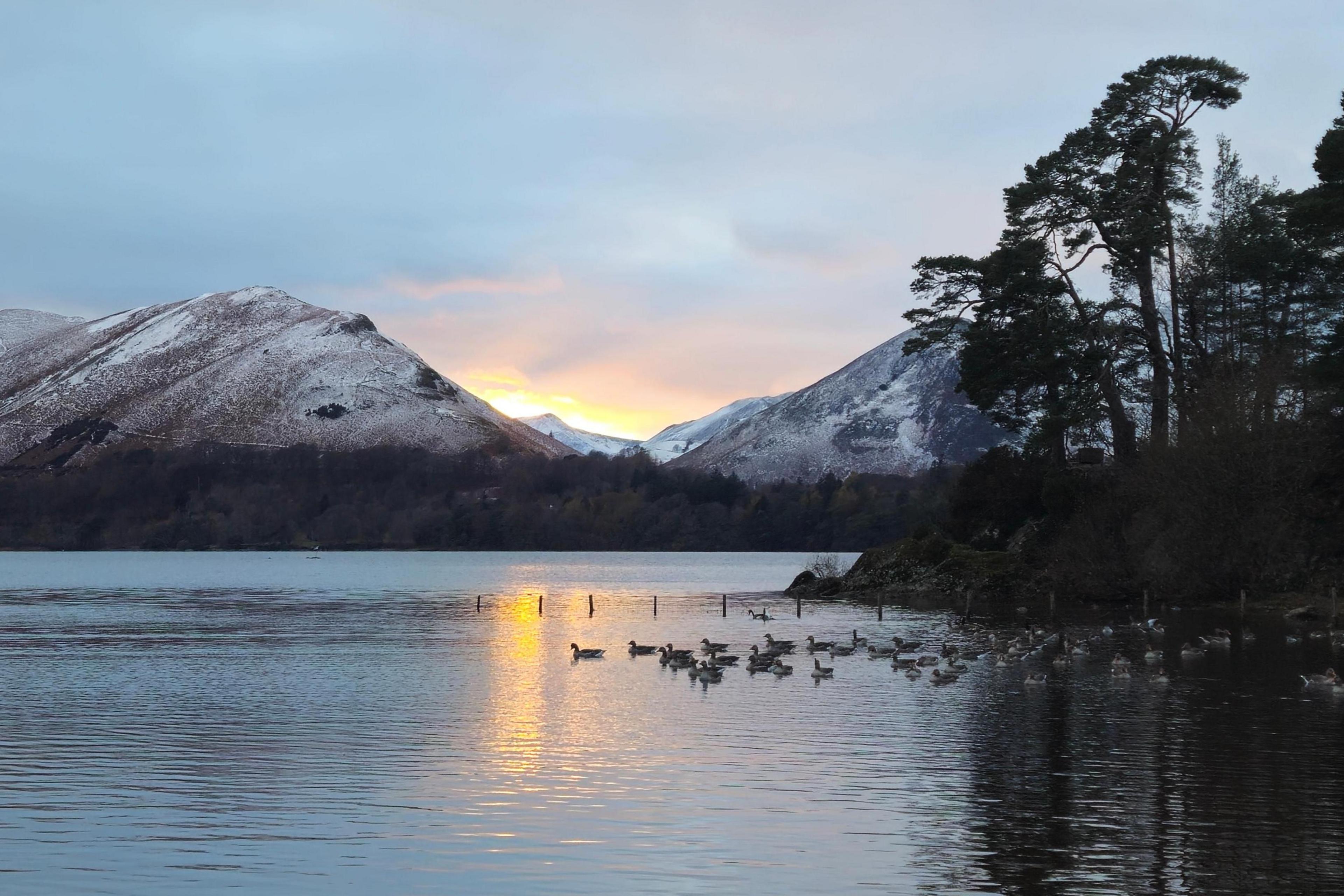The sun is barely poking out from behind the snowy peaks. In the foreground, birds are swimming on a lake which is reflecting the yellow/orange colours of the sky.