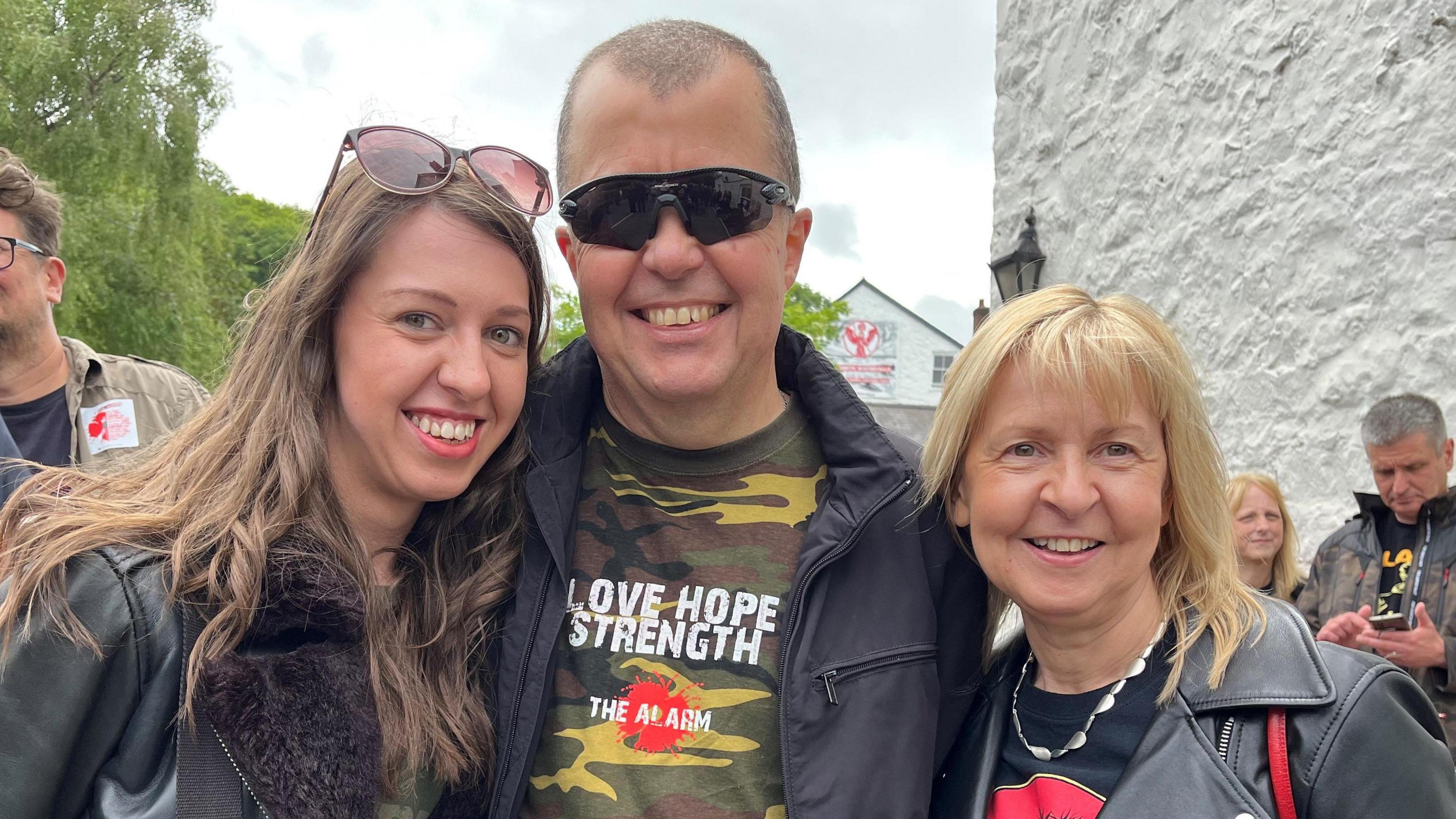 Three people smile at the camera. One women with dark hair, one with blonde hair and a man with short hair and glasses. They are all wearing leather jackets.
