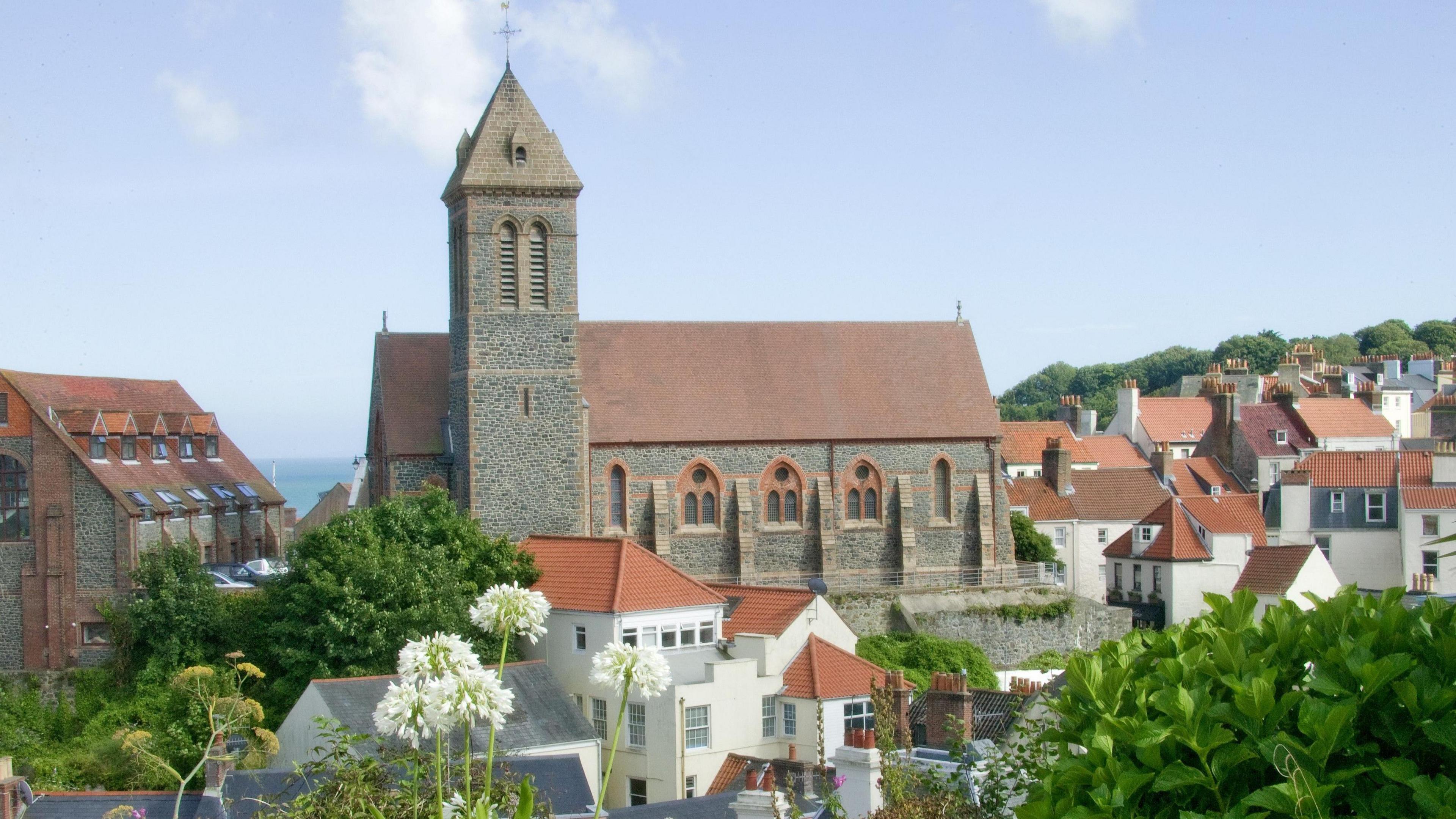 Exterior of a church building. The property is surrounded by smaller residential properties and trees. The sky is blue.