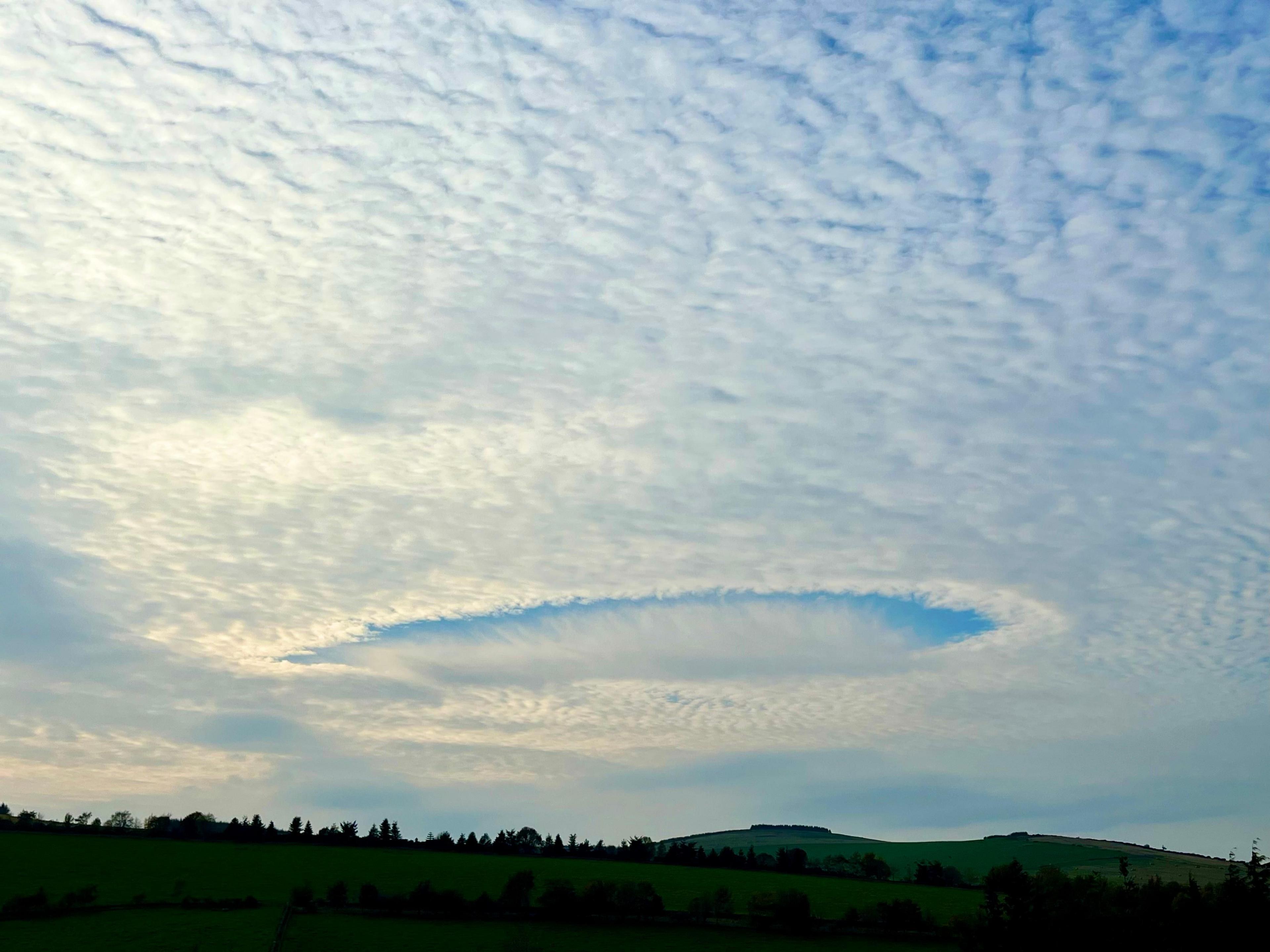 A wide circular hole has formed in a layer of thin white cloud above green fields and gentle hills near Auchleven, Aberdeenshire. The blue sky is visible through the gap, surrounded by textured cloud patterns stretching across the scene.