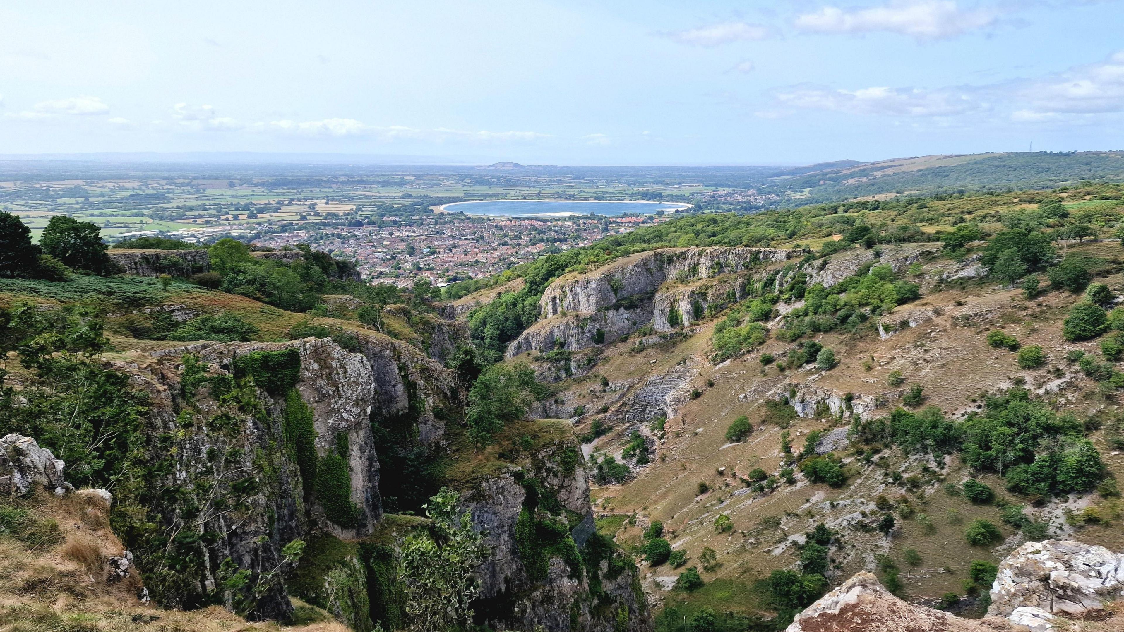 A spectacular view over a valley with cliffs to each side. In the far distance is a circular lake. The sky is blue and the sun is shining.