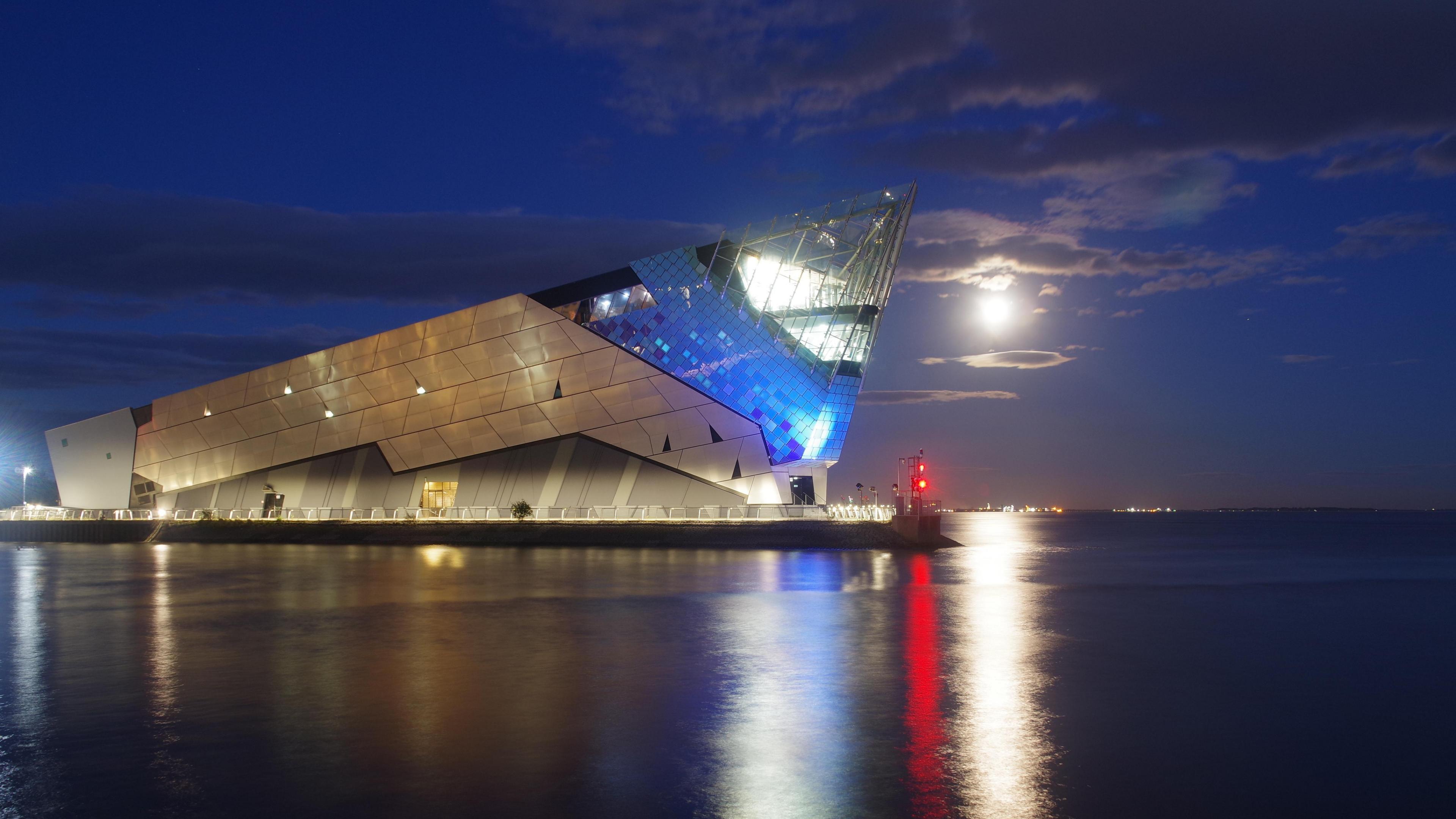 A  sharp angular building sits on a body of water at night time. The building is known locally as The Deep in Hull. It is lit up with glowing blue and white lights that reflect off the water’s surface. Above, a full Moon glows in a dark blue sky scattered with clouds.