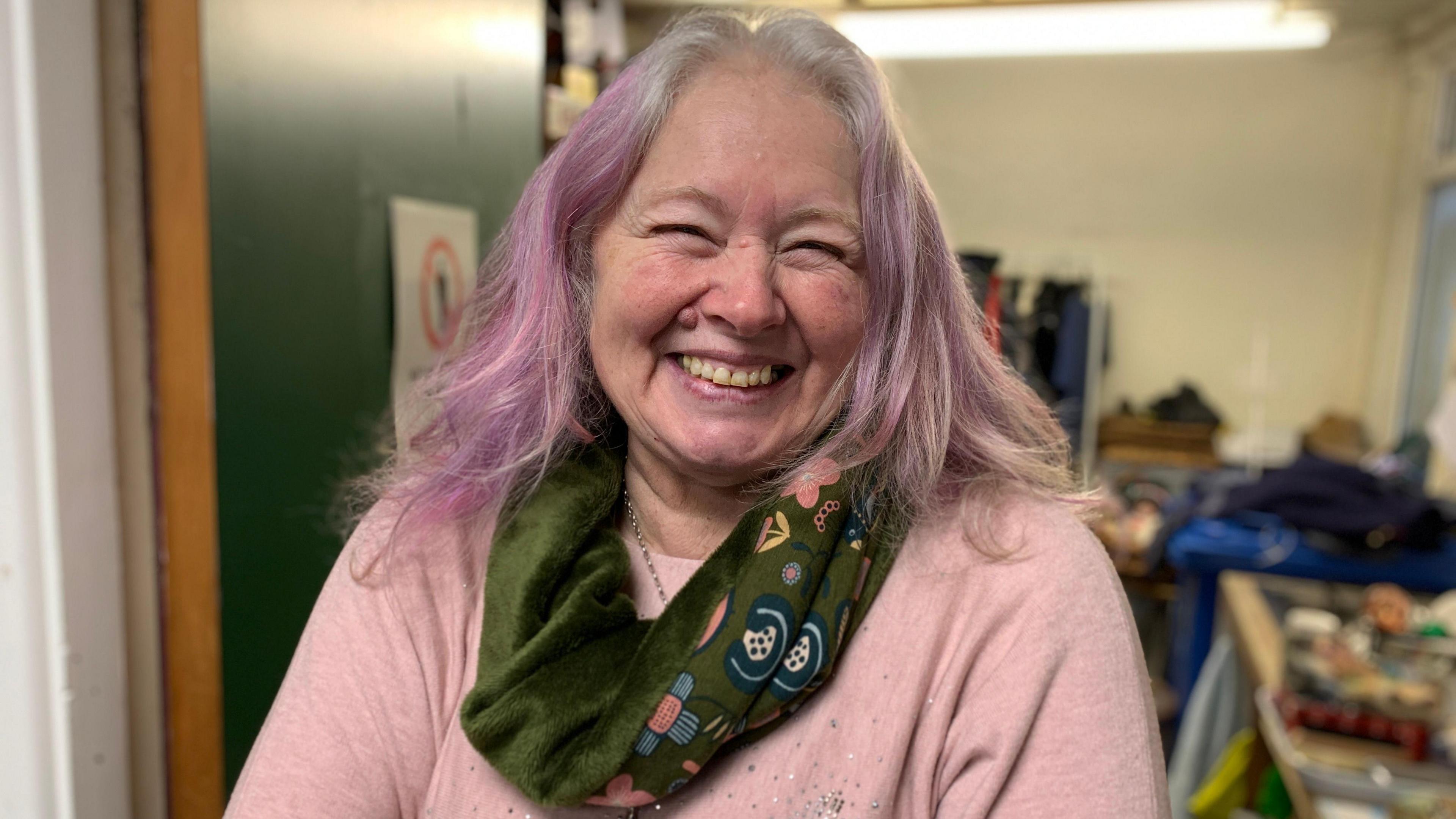 A woman with purple-dyed hair, wearing a pink sweater with a green scarf. She is standing in a shop storeroom doorway. 