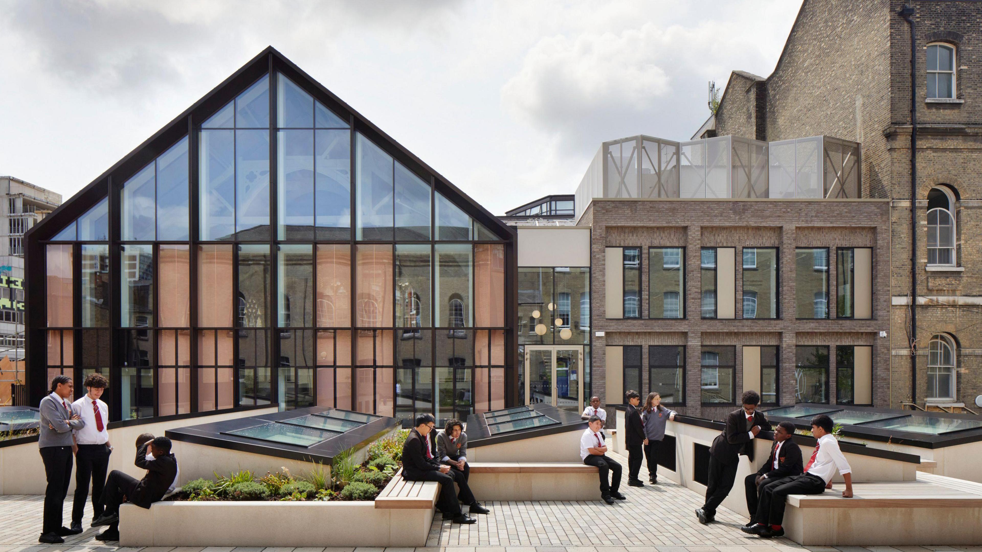 Pupils dotted around a courtyard with a triangular shaped building with a black outline in the background, with lots of vertical windows. There are small shrubs in the courtyard.