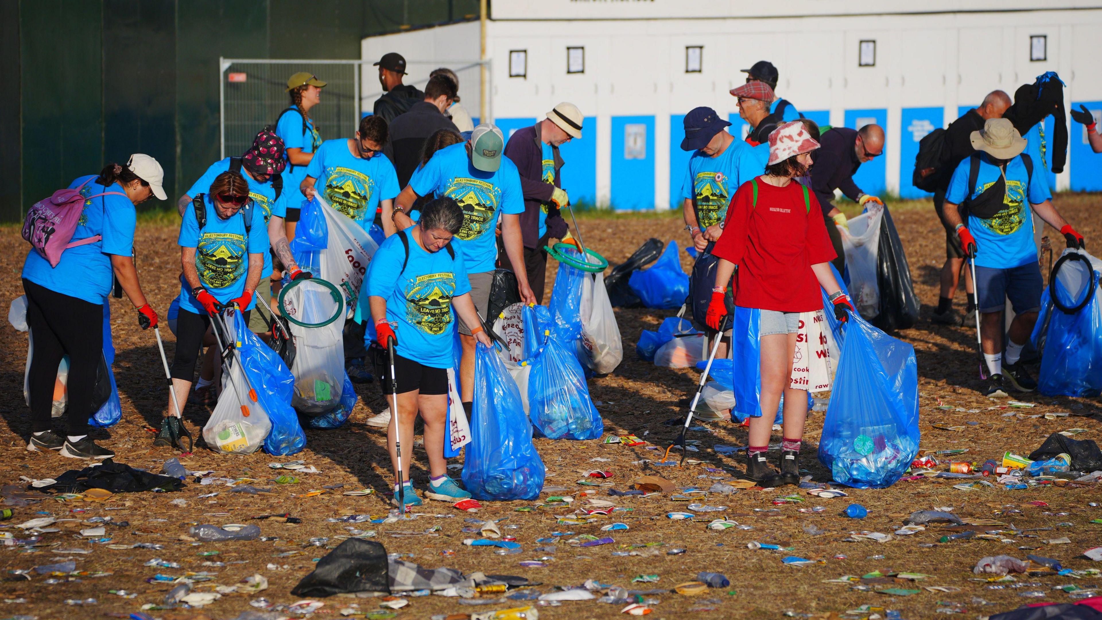 A team of volunteers dressed in blue t-shirts litter picking at Glastonbury. They all have gloves, litter pickers and bags of rubbish.