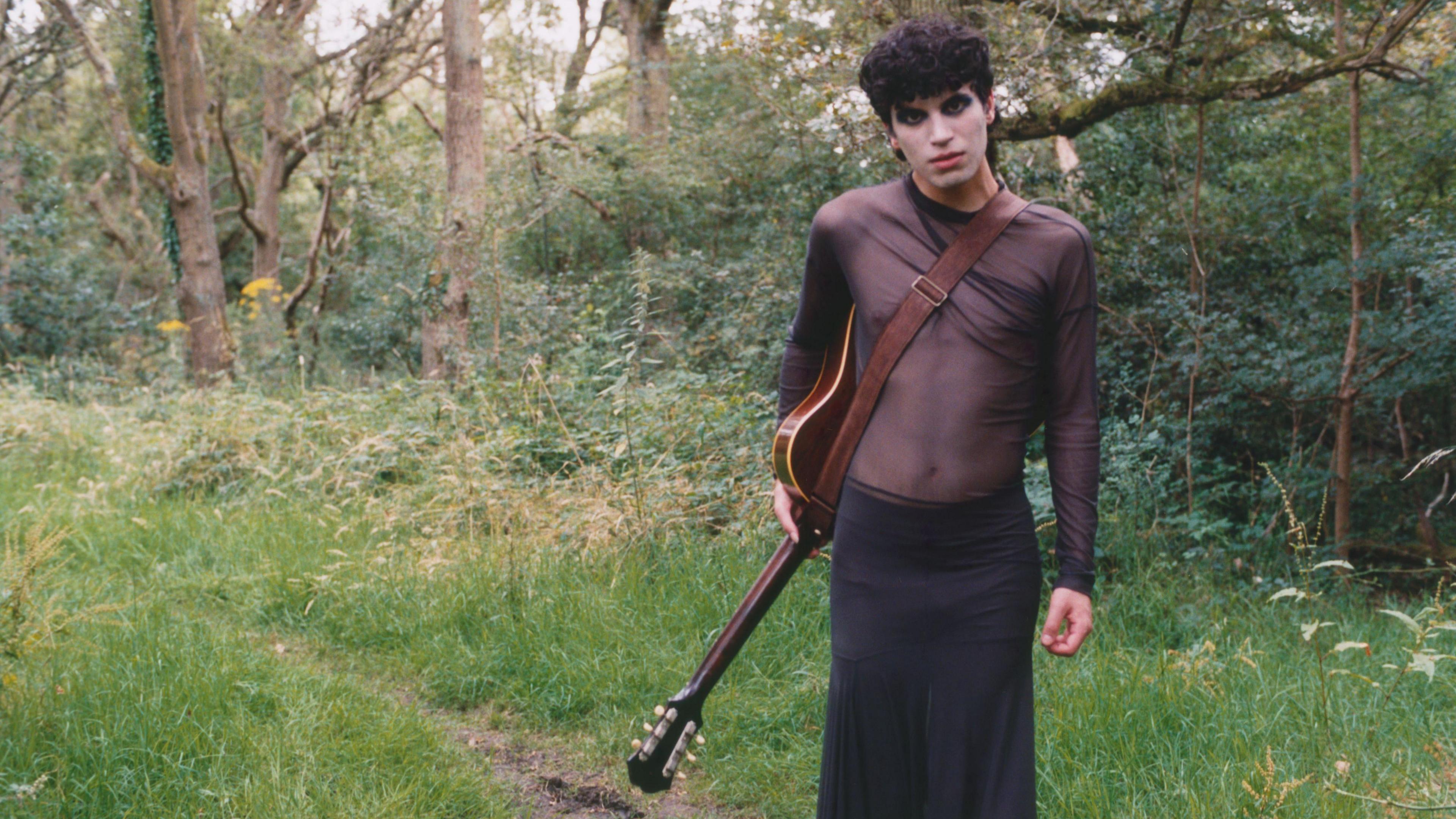 A promo photo of Jacob standing in a forest with an acoustic guitar hanging from their side. They have black hair and black eye makeup. They are wearing a brown, see-through sheer top and a long black skirt.