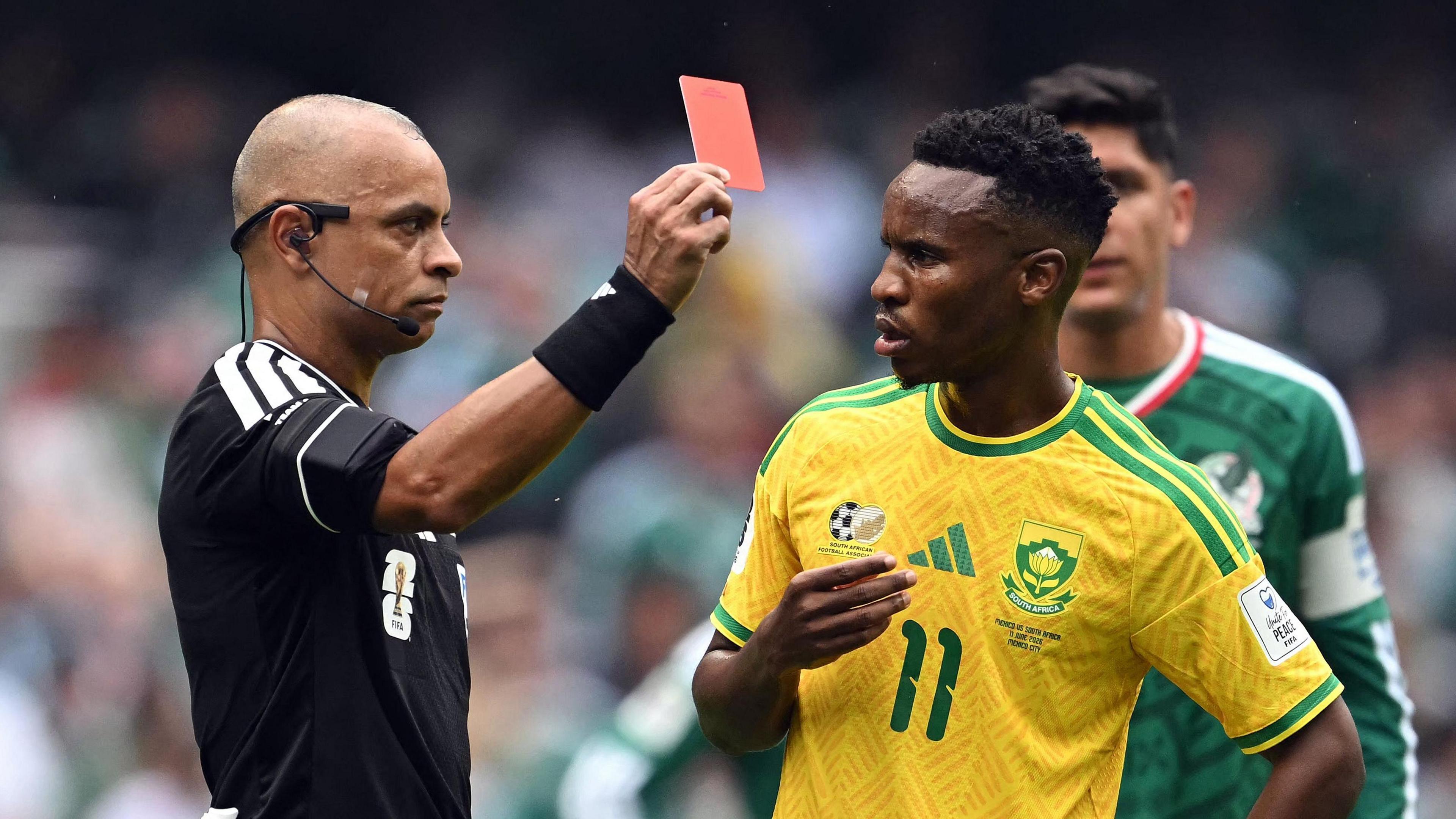 Referee Wilton Sampaio, wearing a black shirt and black sweatband on his wrist, holds a red card with his right hand in front of Themba Zwane, who looks uncomprehendingly at the official while wearing a yellow South Africa shirt with green trim and a green number 11 on the chest. Both men are visible from waist up. A Mexico player in a green shirt is half obscured in the background, with a crowd visible out of focus in the distance