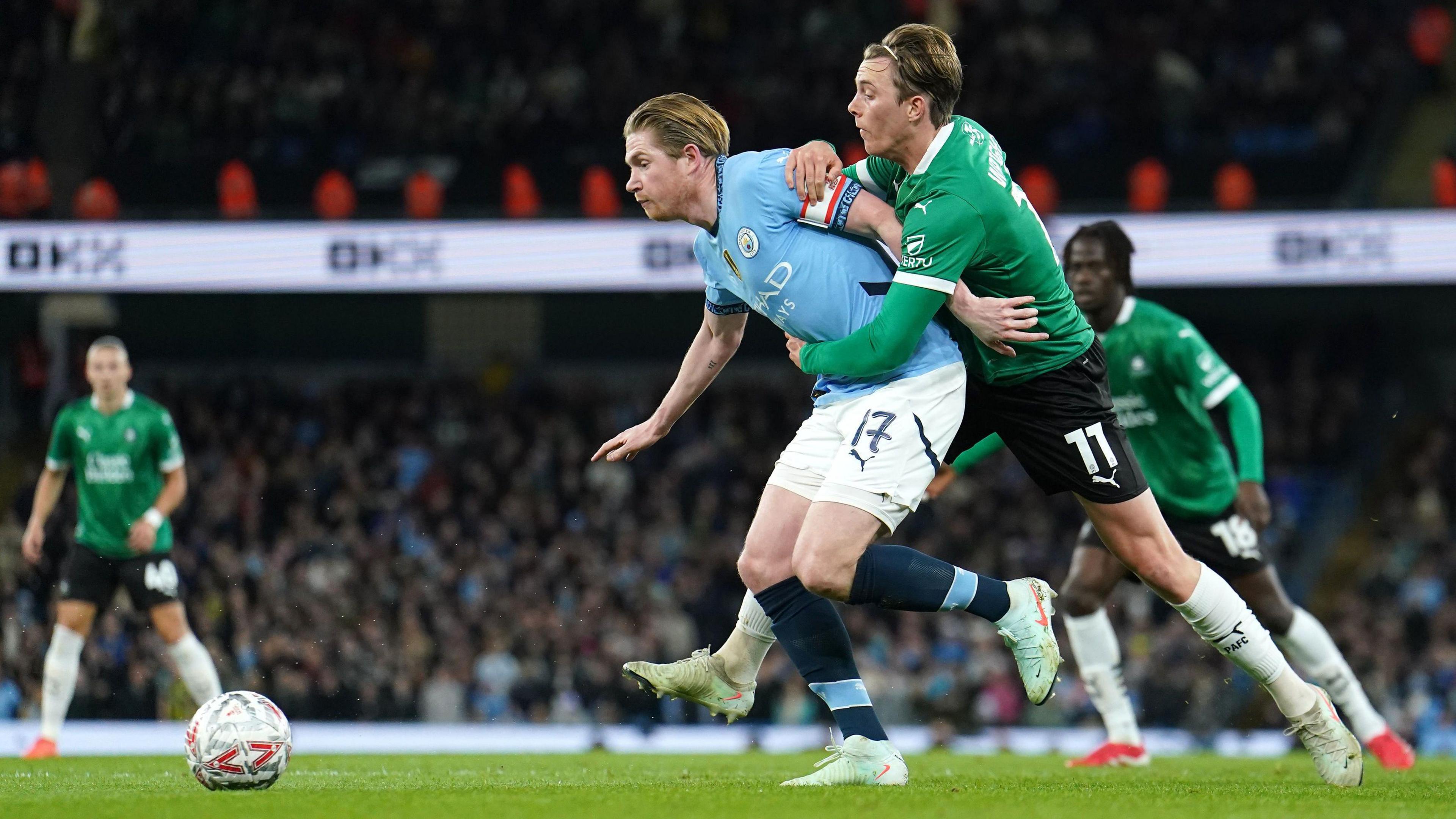 Manchester City captain Kevin de Bruyne and Plymouth Argyle midfielder Callum Wright tussle while fighting to get the ball in the sides' FA Cup fifth round clash at the Etihad Stadium. De Bruyne has a white and red captain's armband on his left arm. Wright is standing behind De Bruyne and is holding onto the City player.