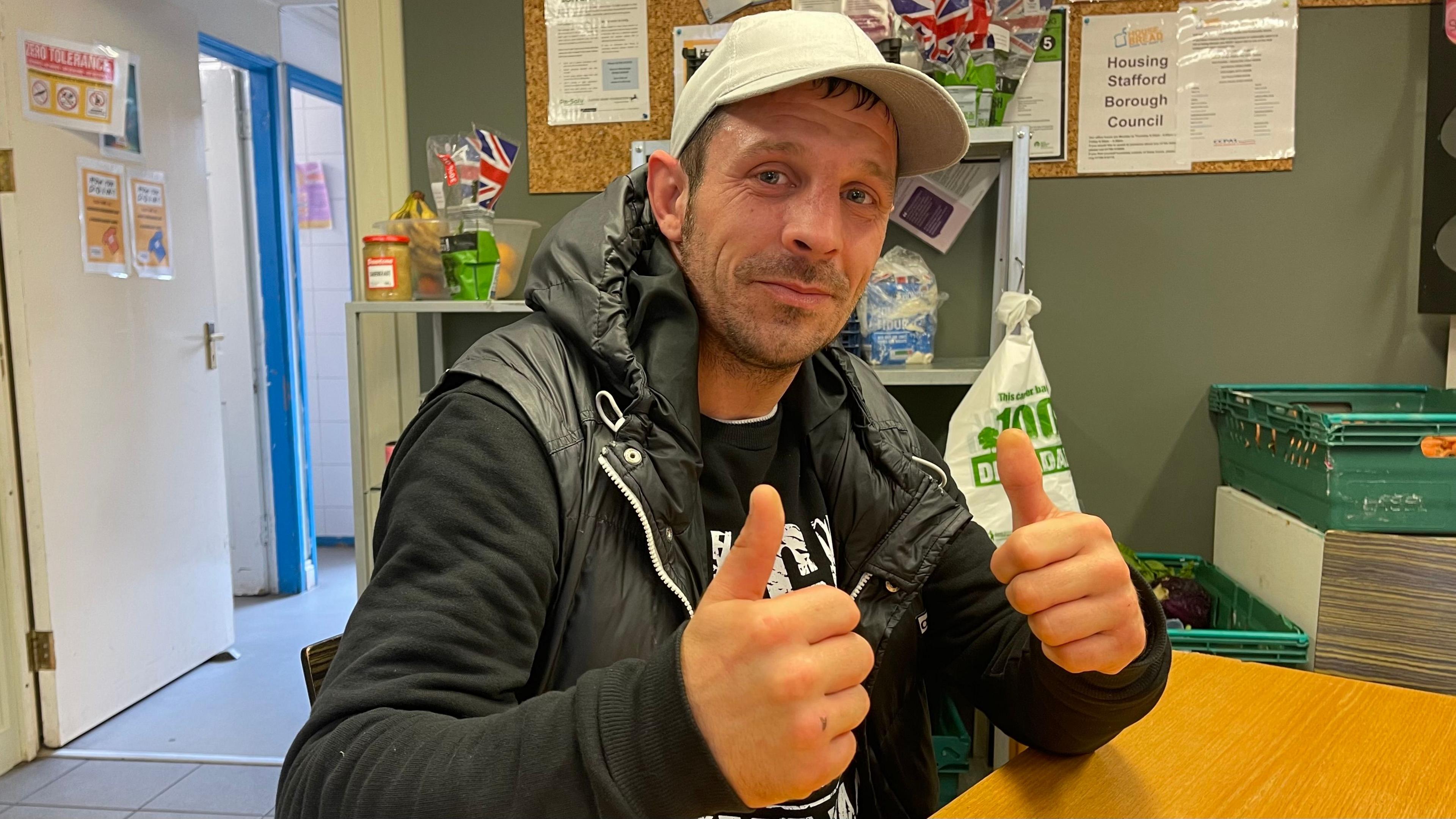 A man in a dark hooded jacket and grey baseball cap sits at a table putting his thumbs up. Behind him are shelves and crates of food.