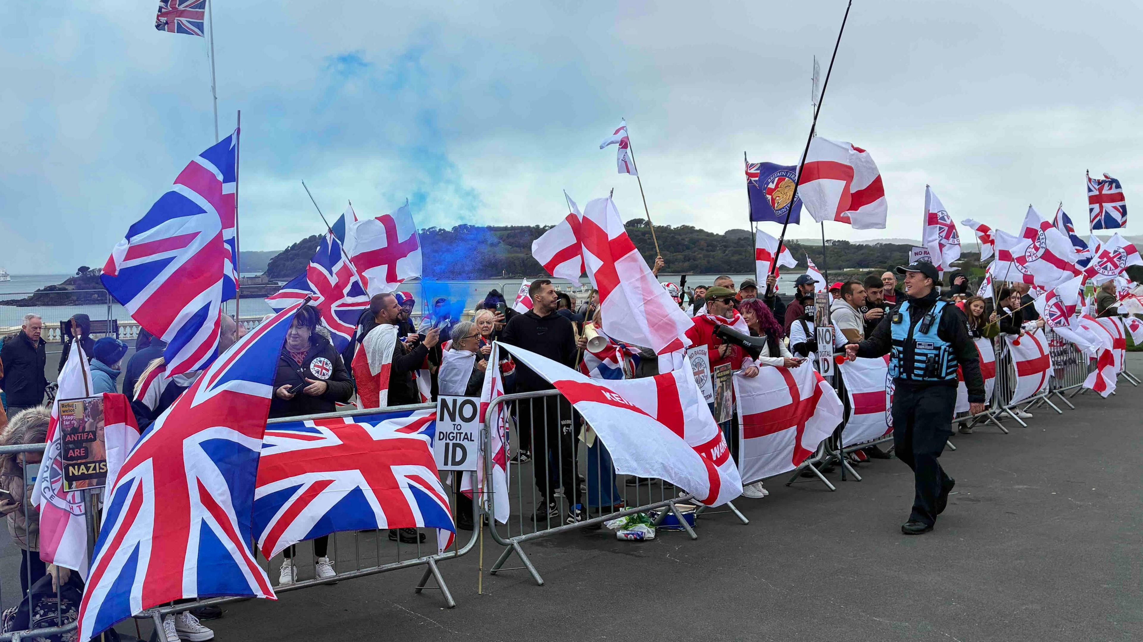 A group of protestors on Plymouth Hoe behind railings waving the Union Jack and St George's flags. A blue flare has been set off and police watch the scene.