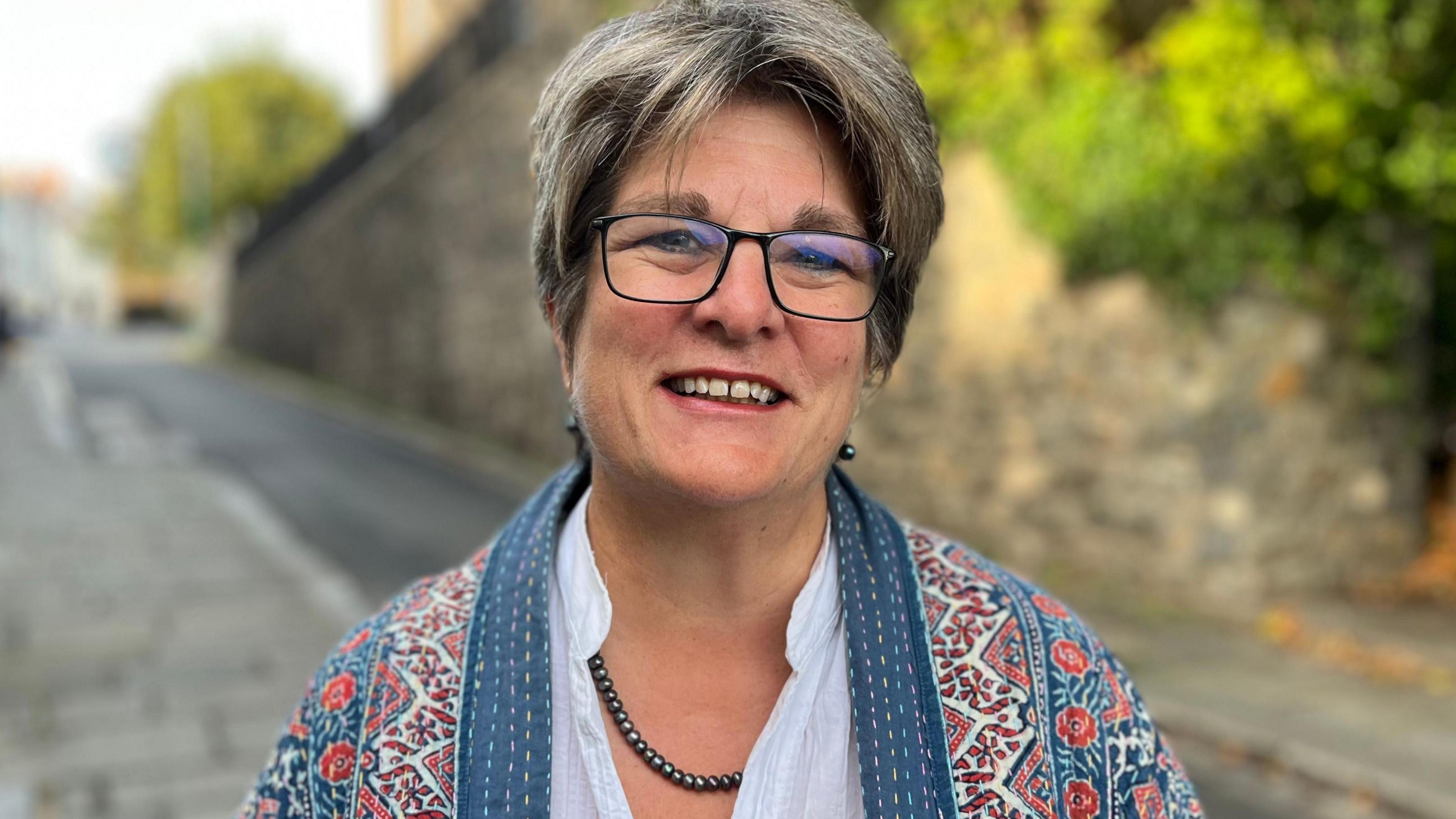 Deputy Jayne Ozanne - A woman with grey hair, black glasses wearing a patterned jacket and a white shirt.