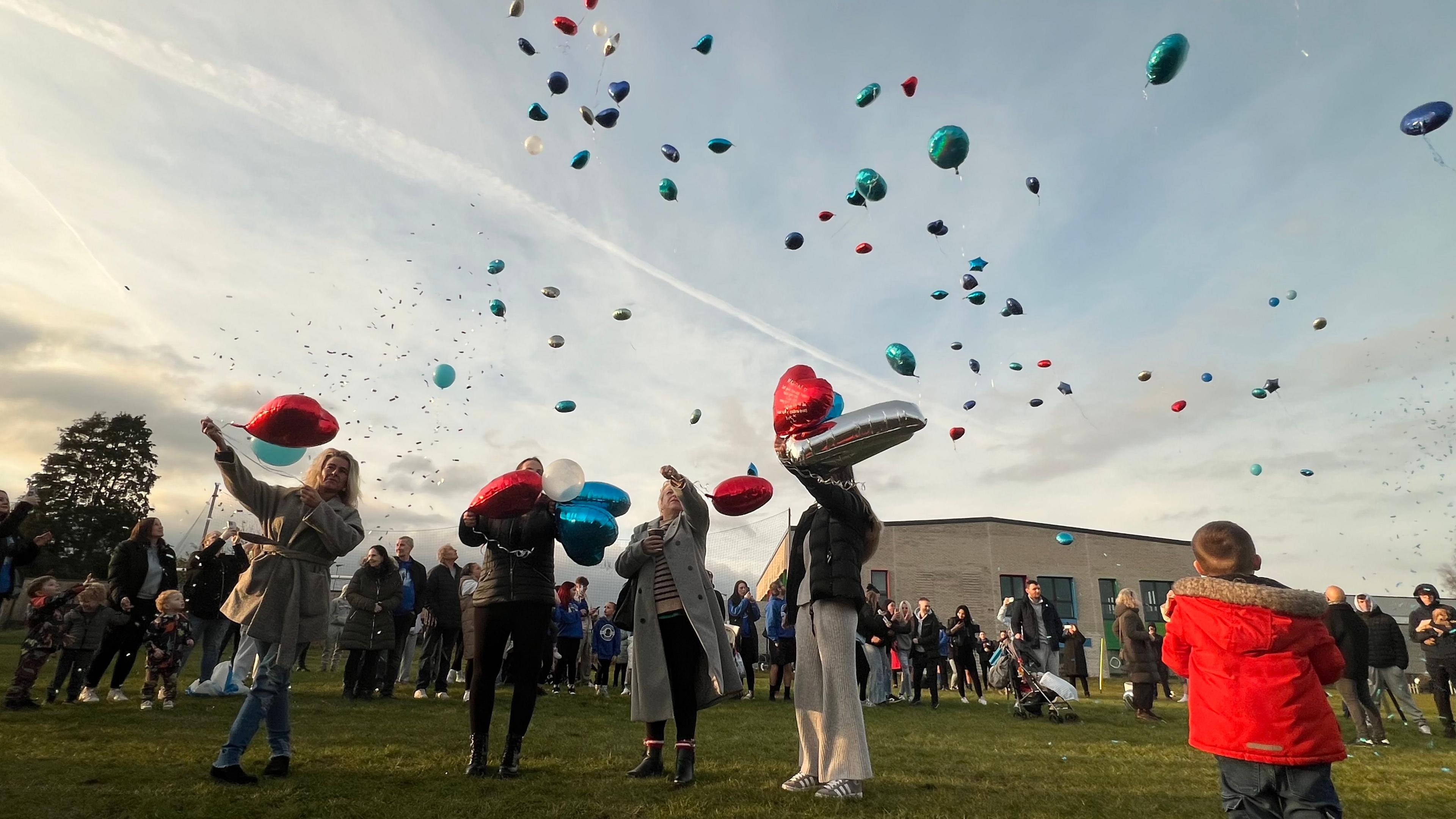 A large crowd gathers in the field of a community centre as they let balloons - mostly red, silver, and blue - into the sky at dusk on a February evening