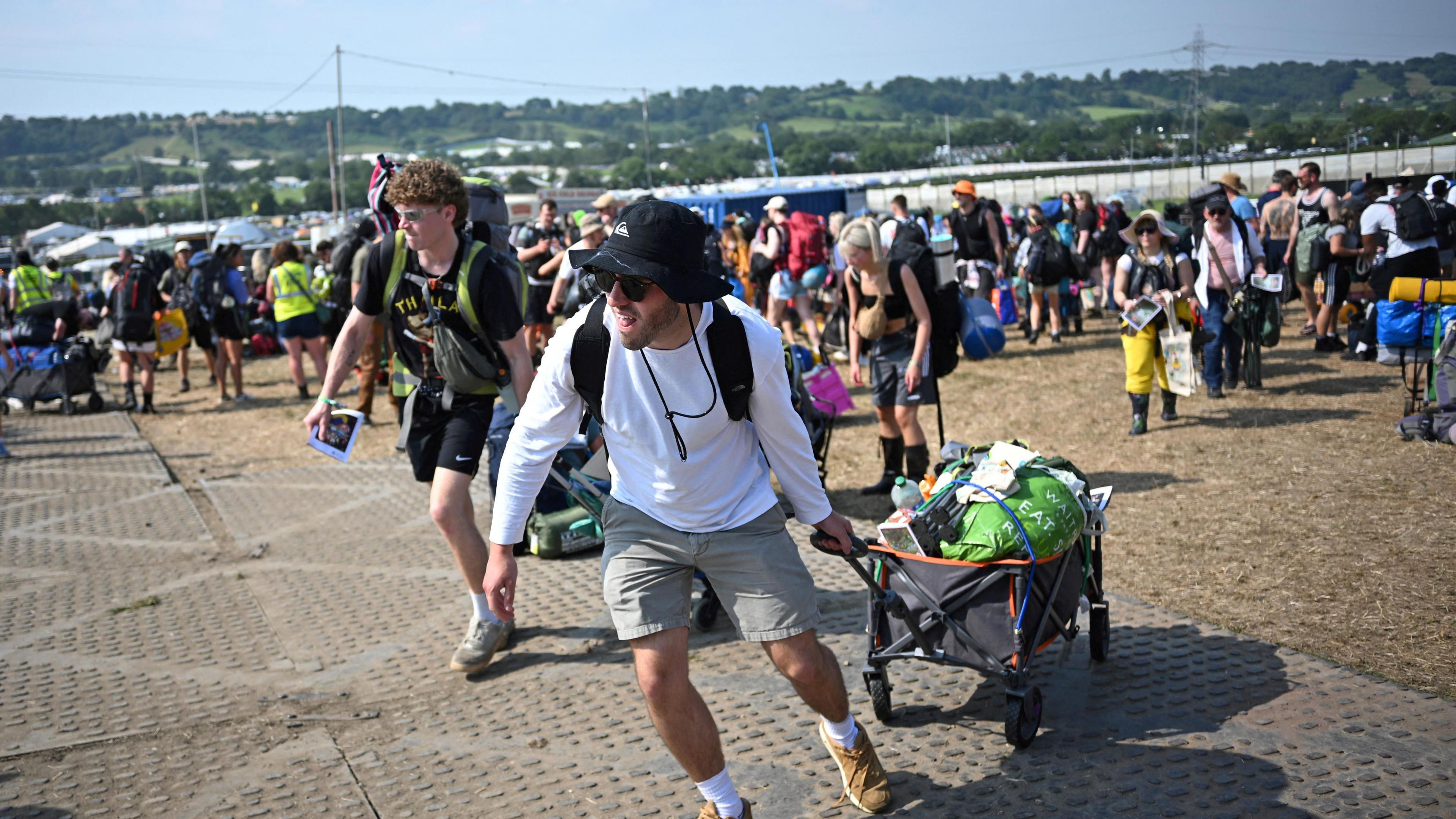 Two young men drag luggage trolleys across the ground at Glastonbury as the sun shines overhead