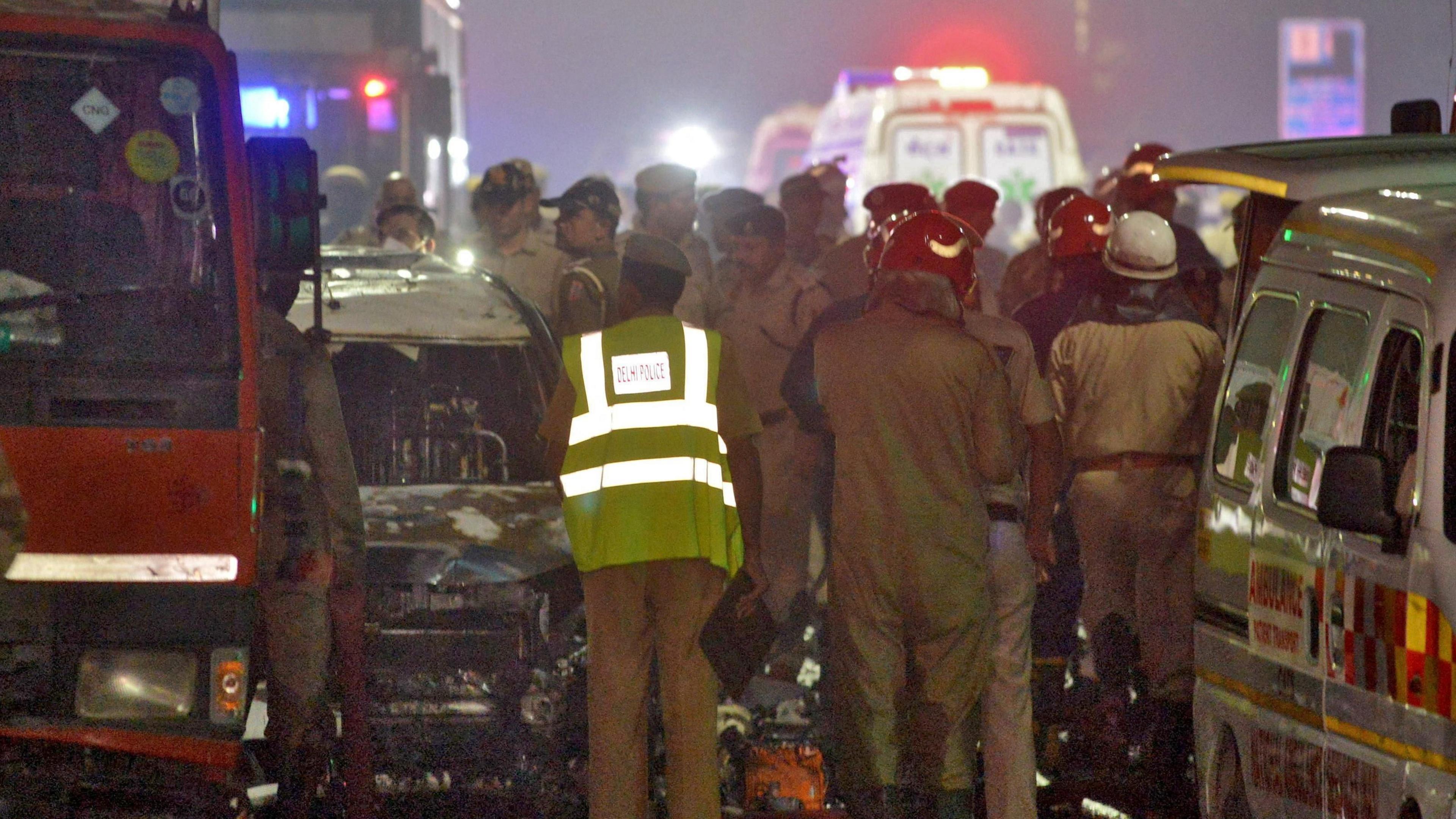 Fire service and other emergency personnel stand next to the exploded car, with ambulances with their lights flashing can be seen in the background