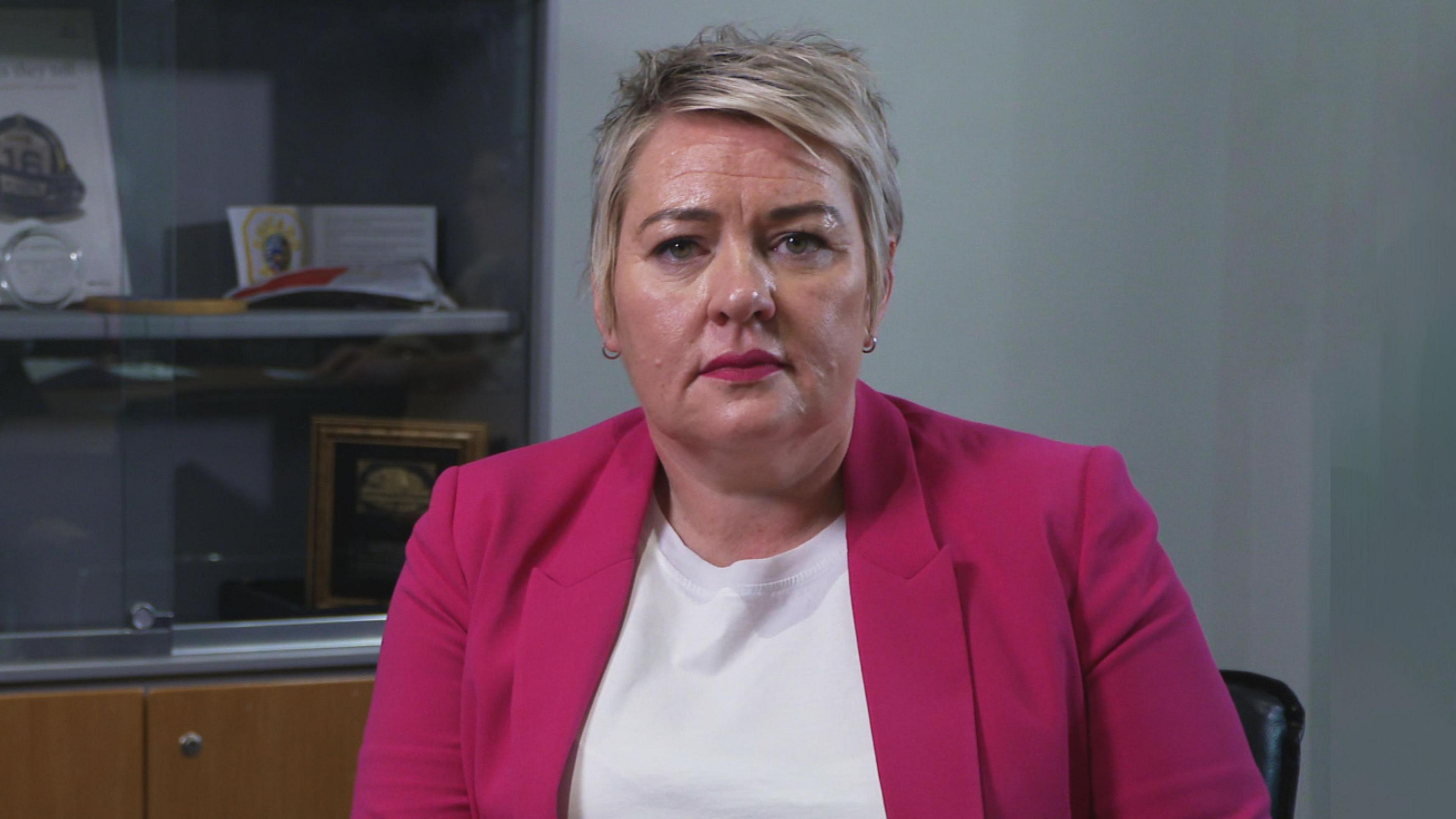 A brown-eyed woman with short cut blonde hair looks directly at the camera. She is wearing a white t-shirt and pink blazer, with two gold hooped earrings in. The woman is sitting in a black chair in a dimly lit grey office. Behind her is a cabinet with a glass door, showing policing plaques and memorabilia.