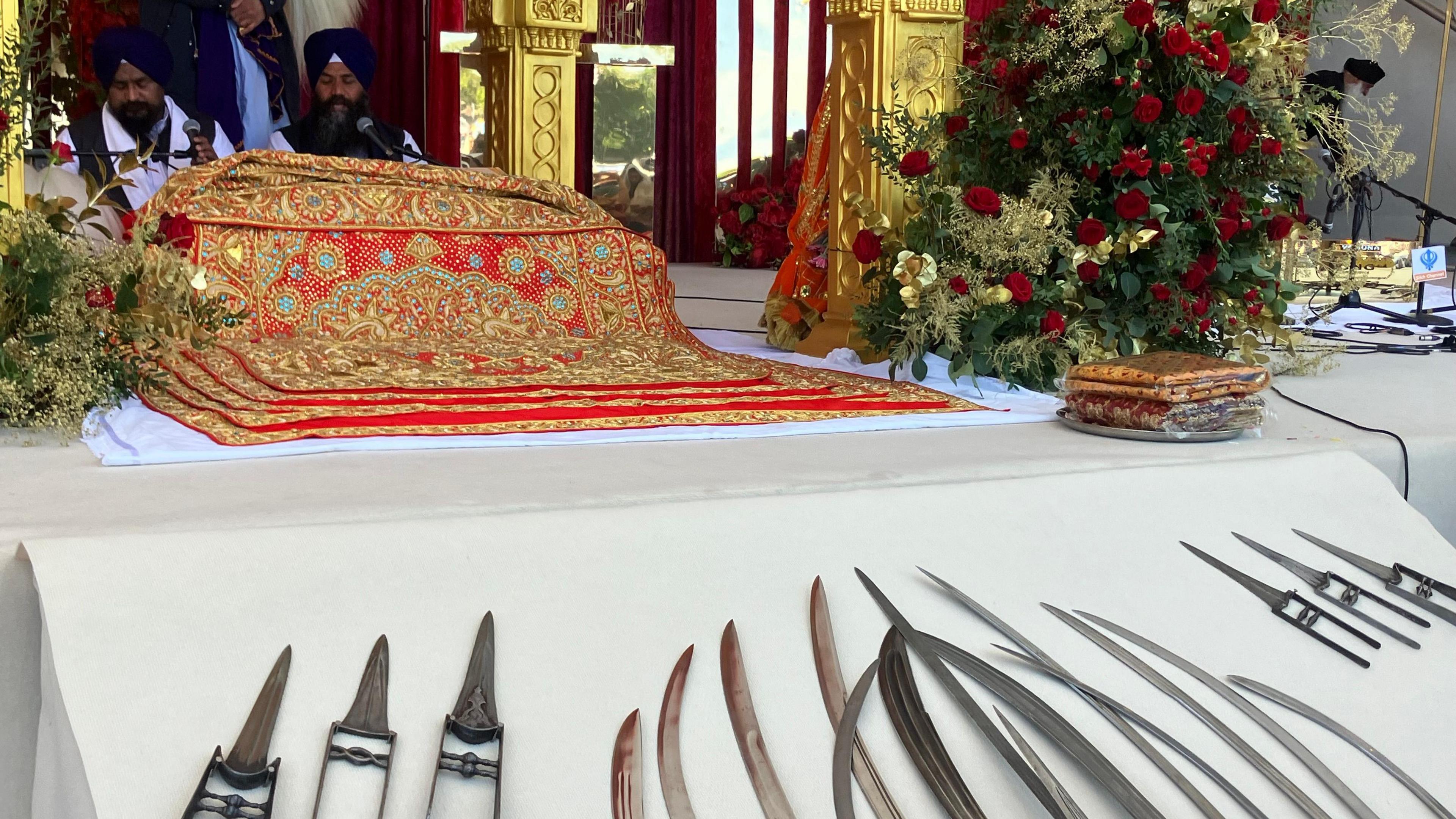 A colourful piece of  cloth covers something on a table covered with a white tablecloth. There is a red and white flower arrangement and inthe foreground there appears to be metal knives. Two men wearing blue turbans are sitting in the background.