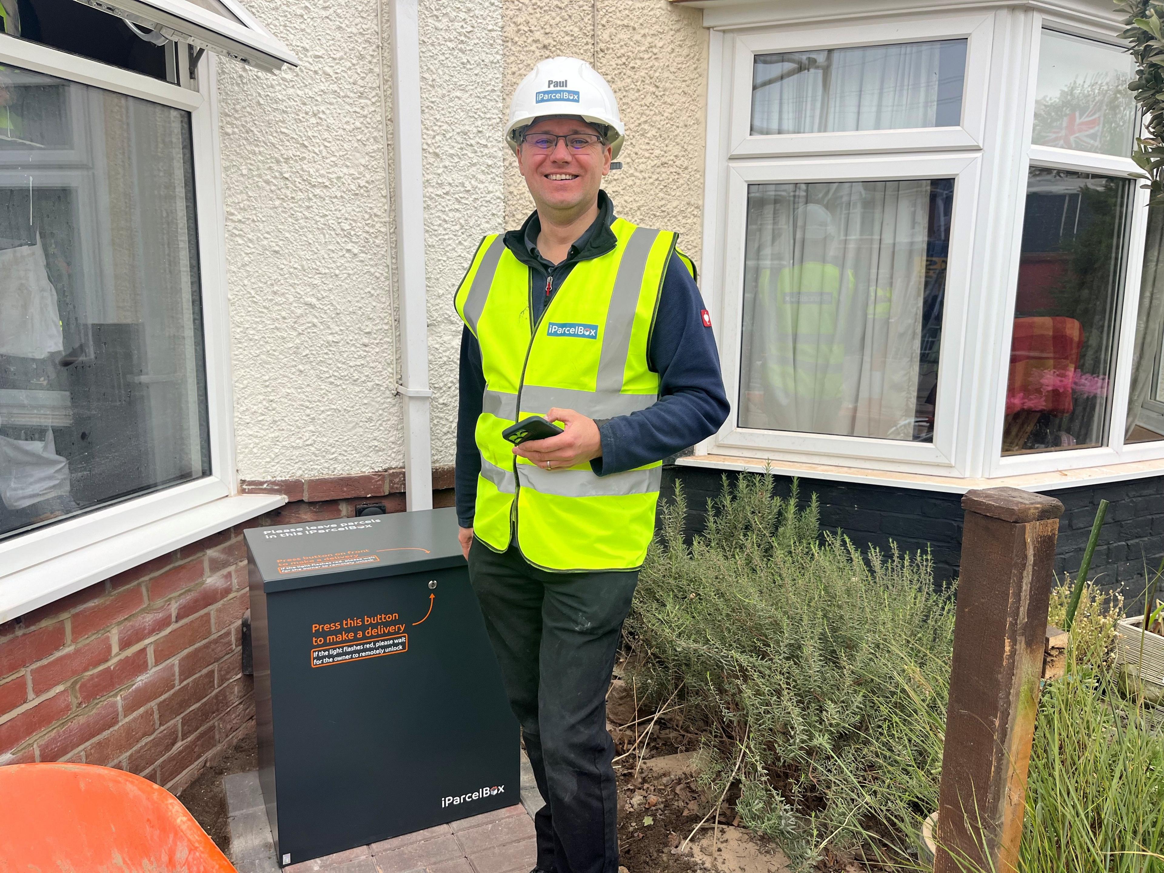 A man wearing a florescent vest and white hard hat standing outside a home next to a large grey box. He is smiling and holding a phone in his hand.