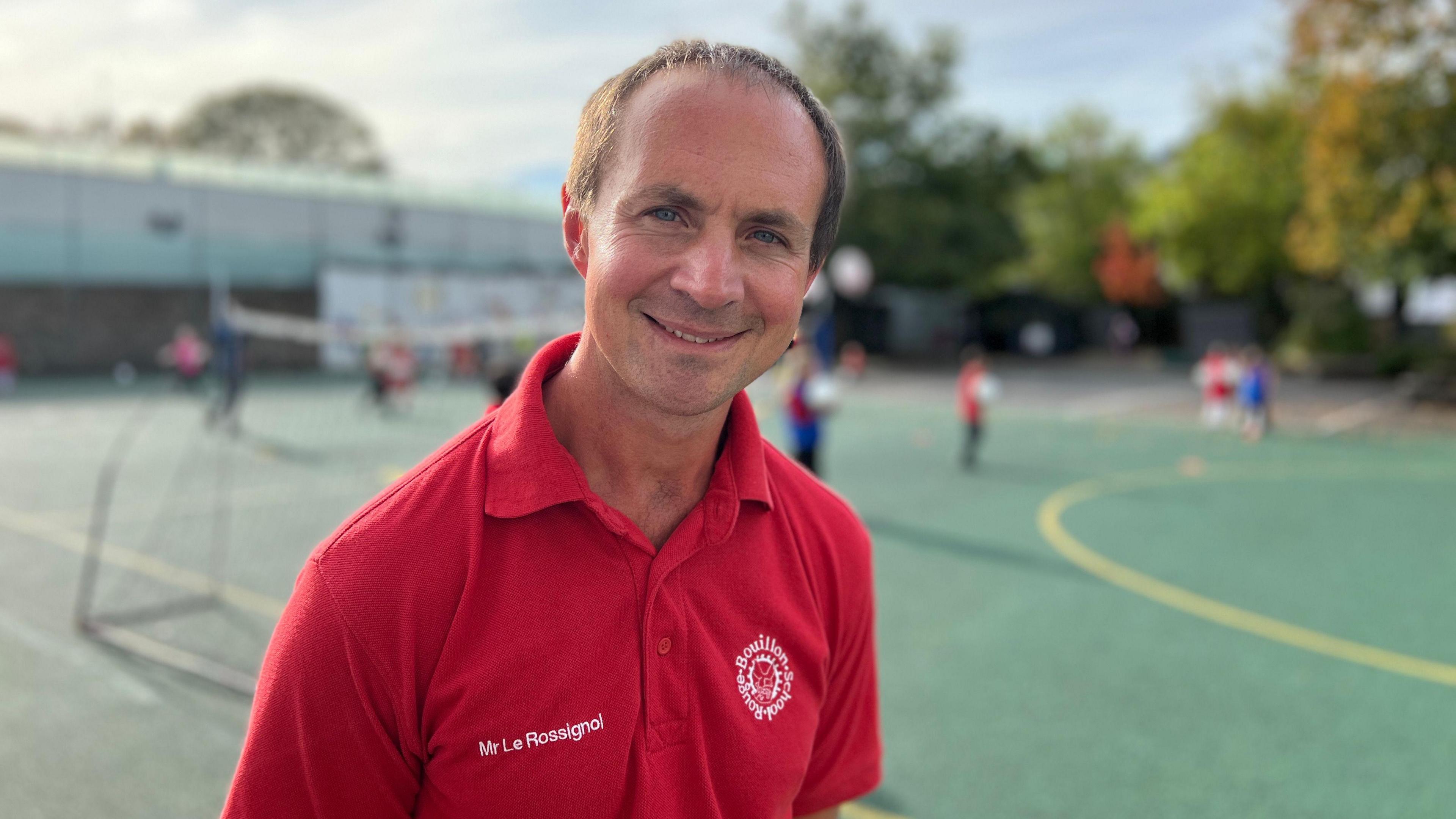 Andre smiles at the camera and he's wearing a red polo top with his name on his chest with the school's logo in white on the other side. Behind him the children are playing football. He has dark hair and blue eyes. His hair line has started to receed.