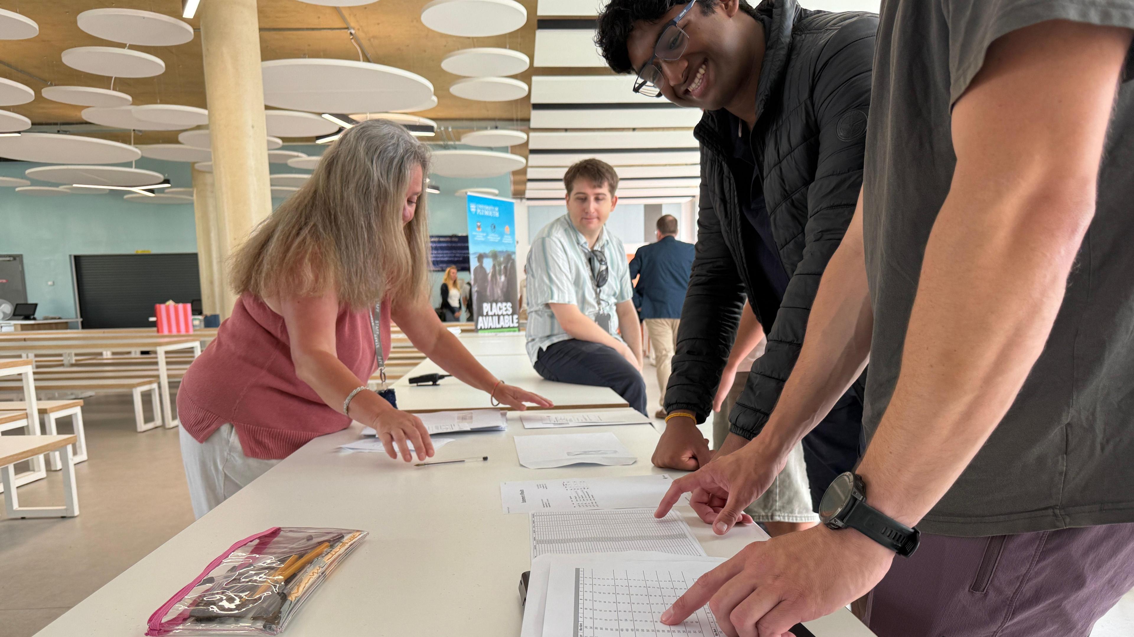A young man is stood waiting to be handed his results. He is leaning his fists on a white table, looking down at a piece of paper, while a man who is slighty out of vision points towards the paper. The young man wears glasses and a dark grey puffer jacket, he is smiling while looking down at the paper. There is a woman with long greying hair on the other side of the table, she wears a pink sleeveless top and white trousers and is moving paper around on the table. In the distance there are more people gathered.
