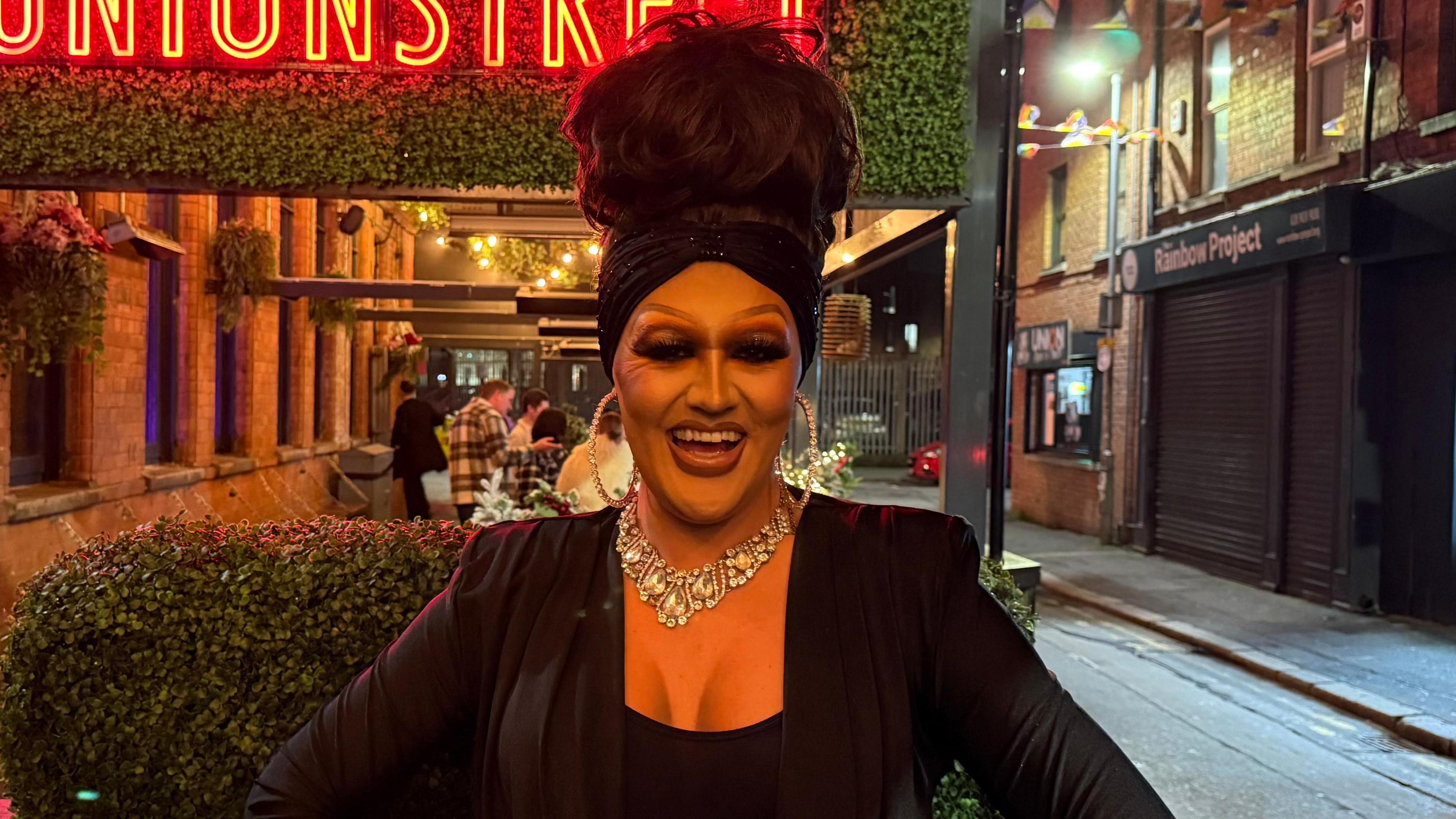 Roxy is standing outside on a street with bright neon signs behind her. She has her long brown hair up in a tall bouffant hairstyle with a turban style hair band. She is wearing full drag makeup and a glitzy necklace over a black fitted blazer.  