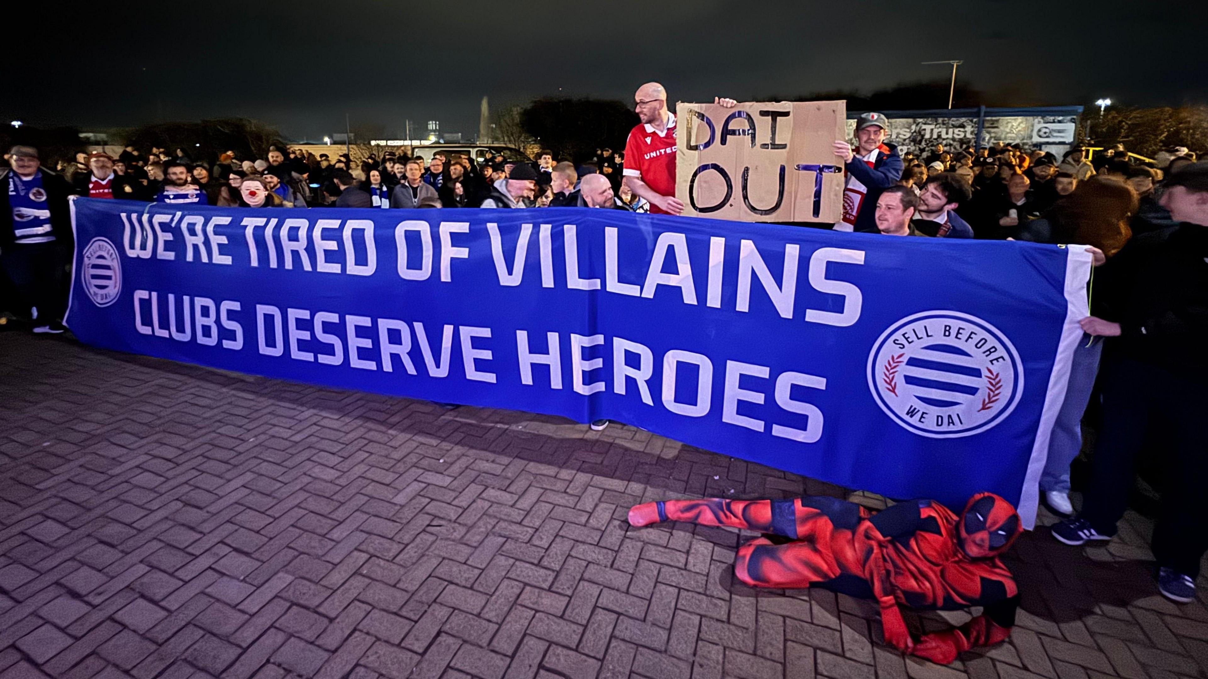 A crowd of people holding up a blue and white banner saying "we're tired of villains, clubs deserve heroes". A man standing behind the banner is also holding up a cardboard sign saying "Dai out", and a person dressed in a red and black Deadpool costume is posting in front of the banner