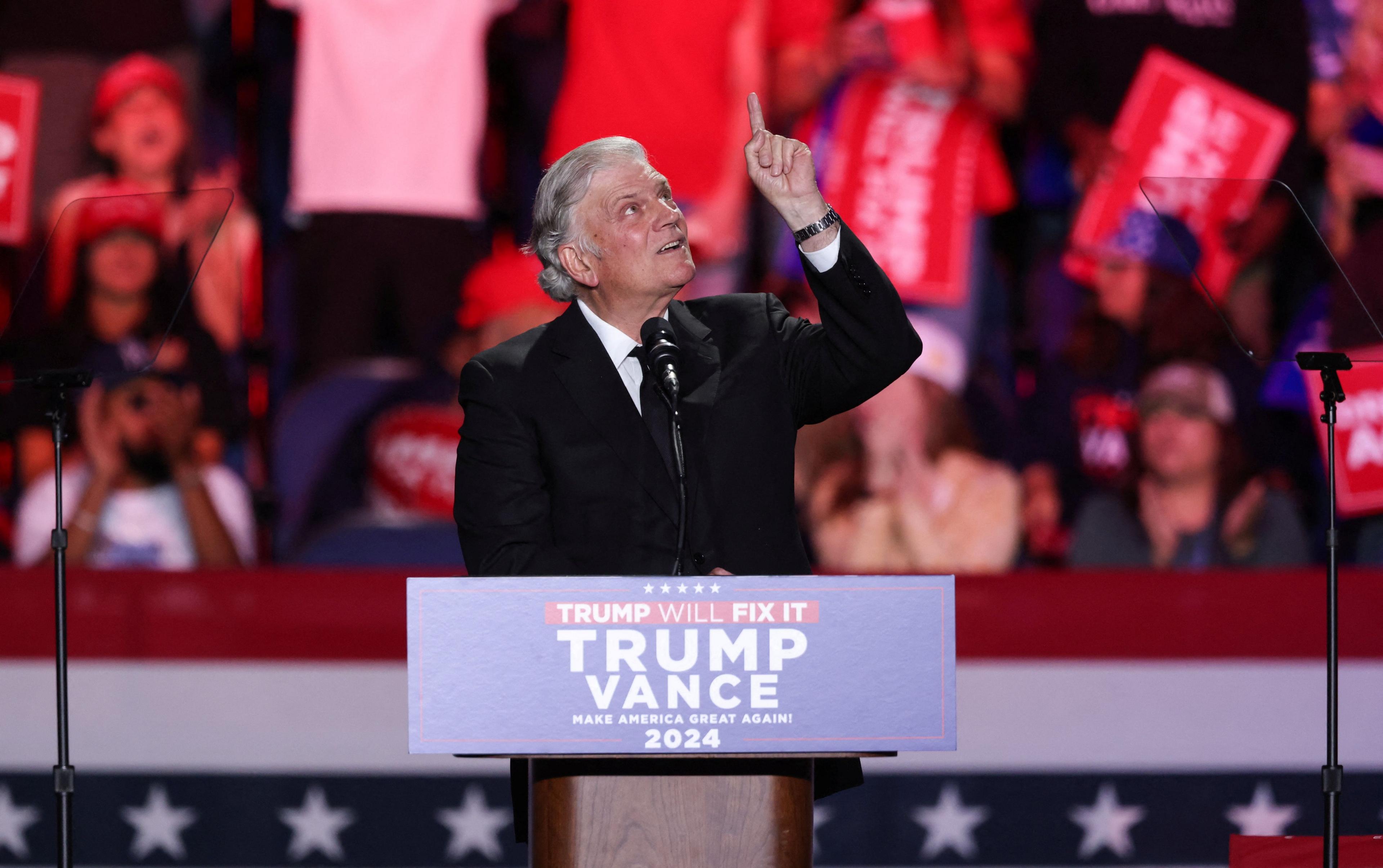 Rev Franklin Graham points upwards whilst standing in front of a Trump Will Fix It stand at a rally