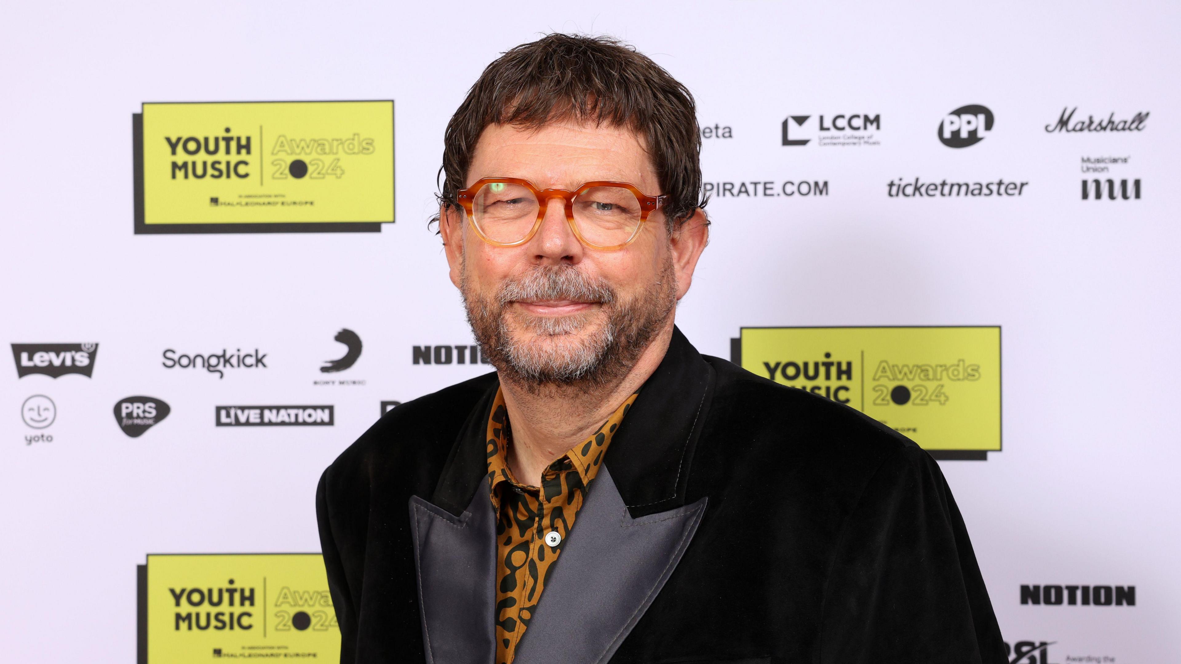 Matt Griffiths at a Youth Music event. He wears a black velvet jacket over a leopard-print shirt, paired with round orange and yellow ombre glasses. 