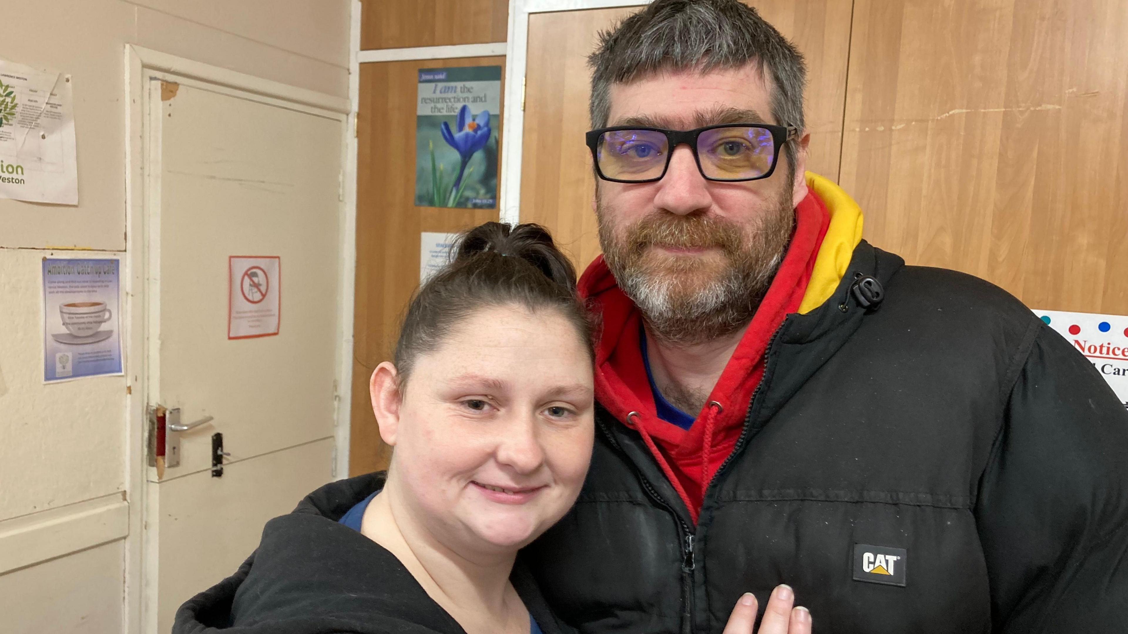 Carole Keene wearing a dark hoodie and top with dark hair and Wayne Harrington wearing a dark coat and a red and yellow hoodie wearing glasses, holding each other and smiling at the camera