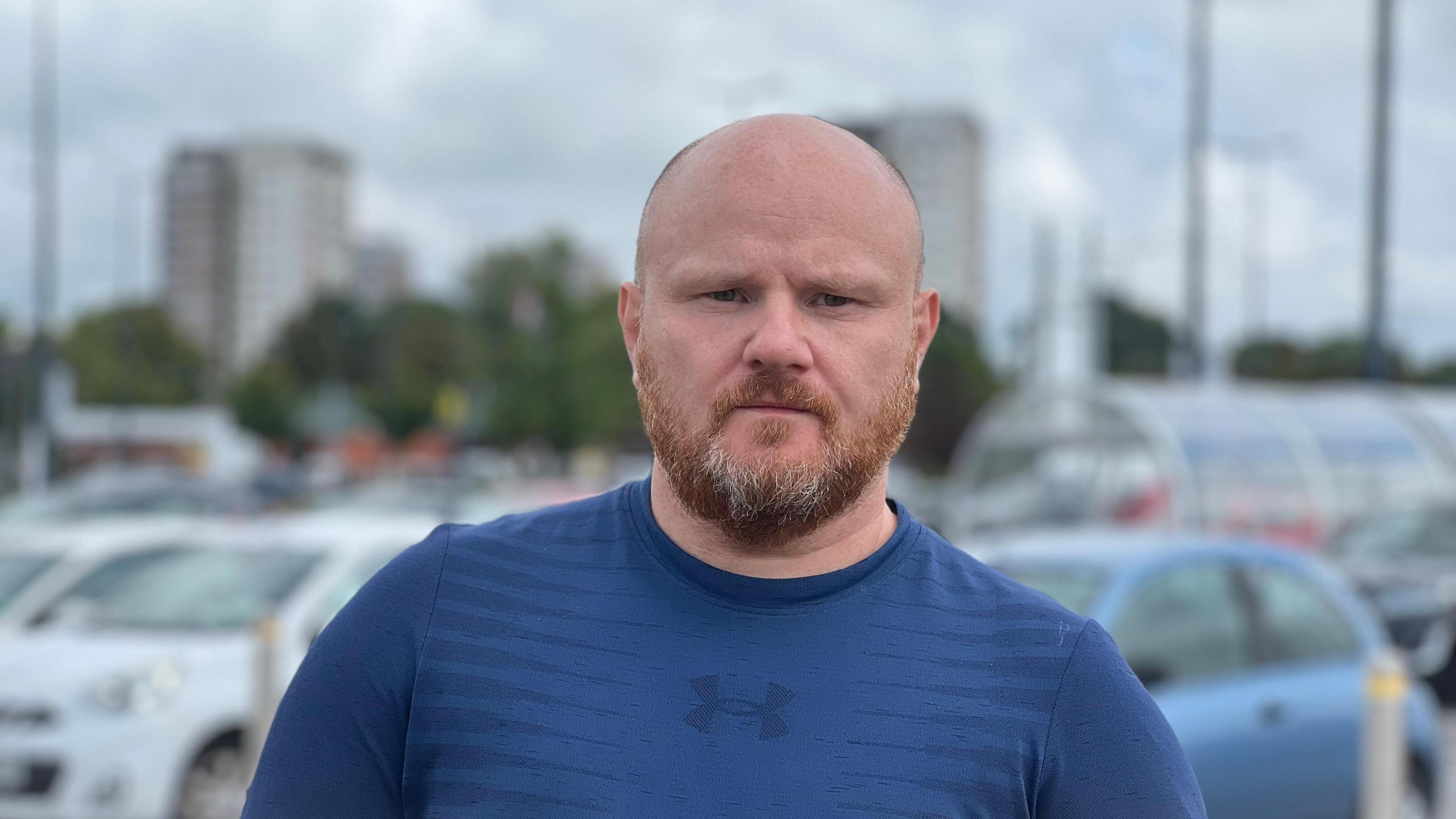 Michael Jones, who has a bald head, short ginger-grey beard and is wearing a blue t-shirt, is standing in front of parked cars with Beech Rise and Willow Rise in the background.