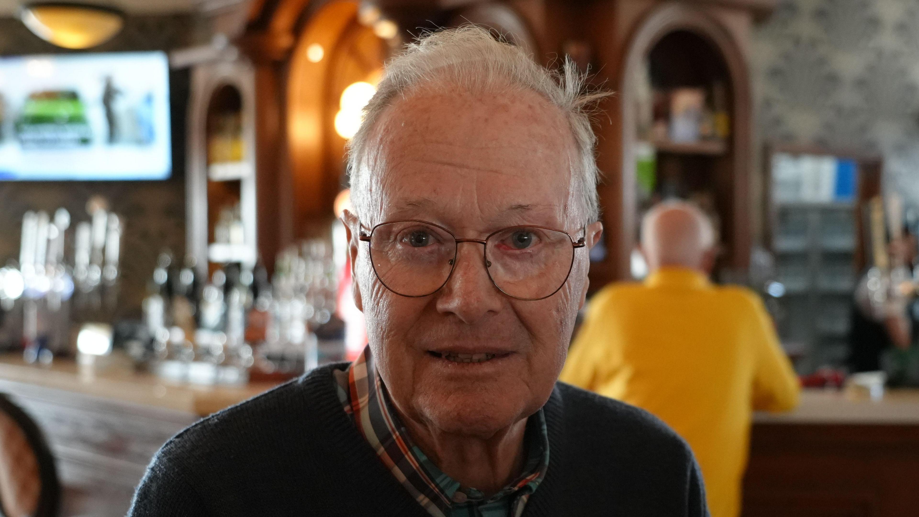 Christopher Lambert looking into the camera. He is wearing glasses and a dark coloured jumper over a shirt. He is in a pub.