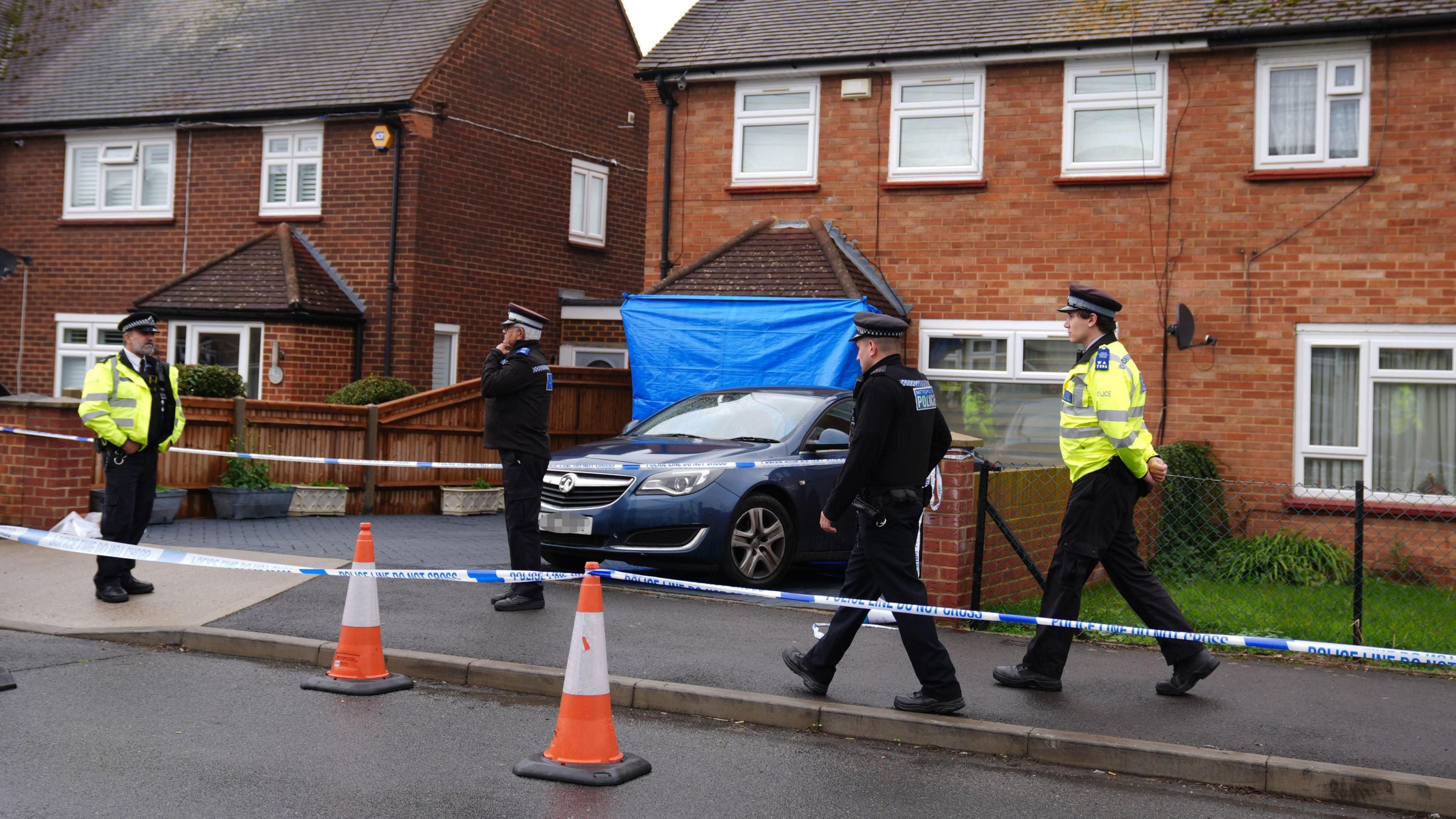 Police officers stand outside a house in Uxbridge, with a blue forensics tent set up in front of a car parked on the driveway. The area is cordoned off with police tape and traffic cones as officers guard the scene.