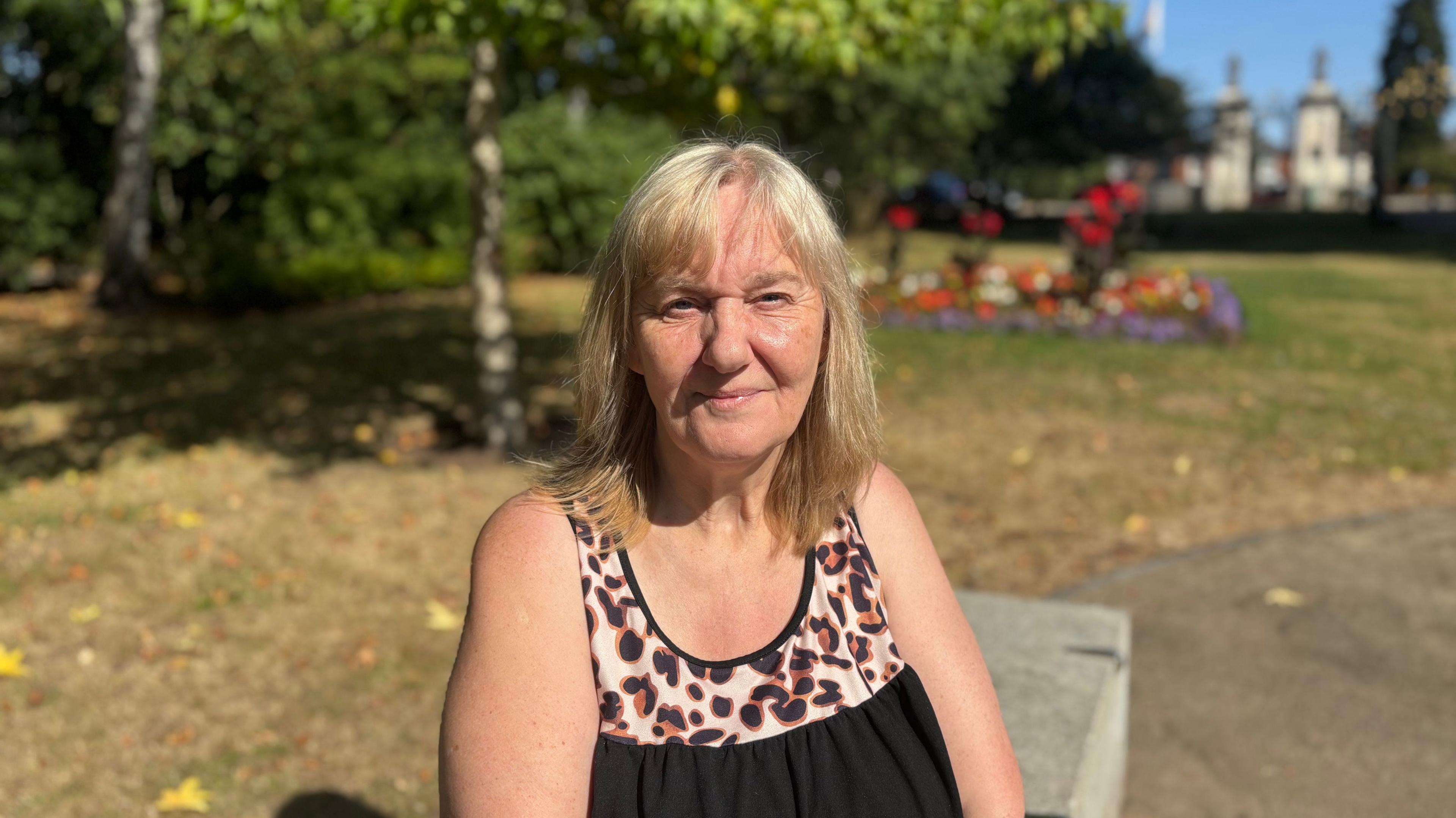 Vi Wood is wearing a black sleeveless top with a leopard-print neckline is seated outdoors in a park or garden setting. Trees, flowerbeds and a pathway are visible in the background, with bright sunshine and clear skies illuminating the scene. The photograph is focused on the person in the foreground, while the surrounding greenery and landscaping are softly blurred.