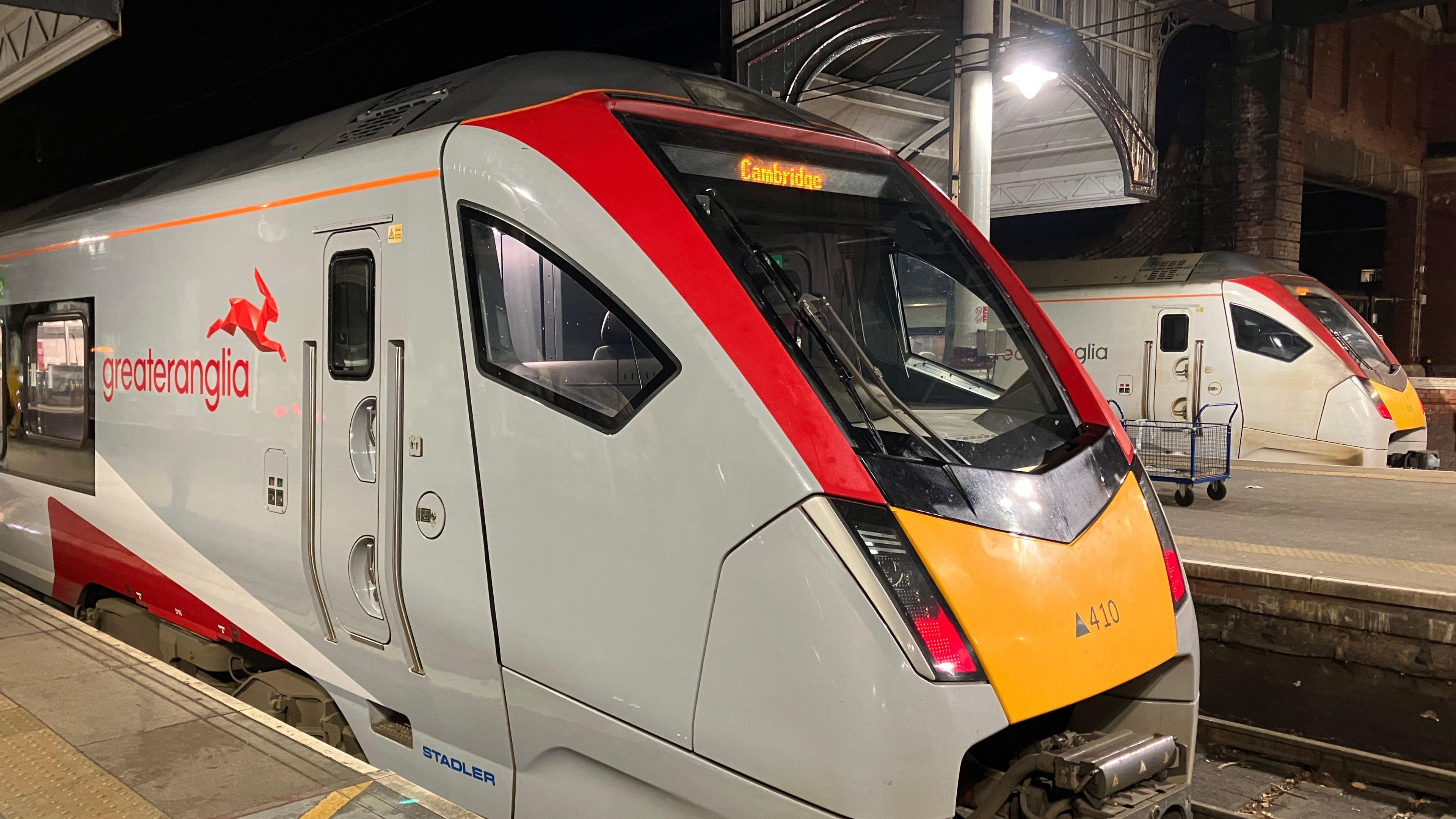 Two grey Greater Anglia trains are shown with yellow and red livery as they are parked at a station's platforms, shown from a side angle.