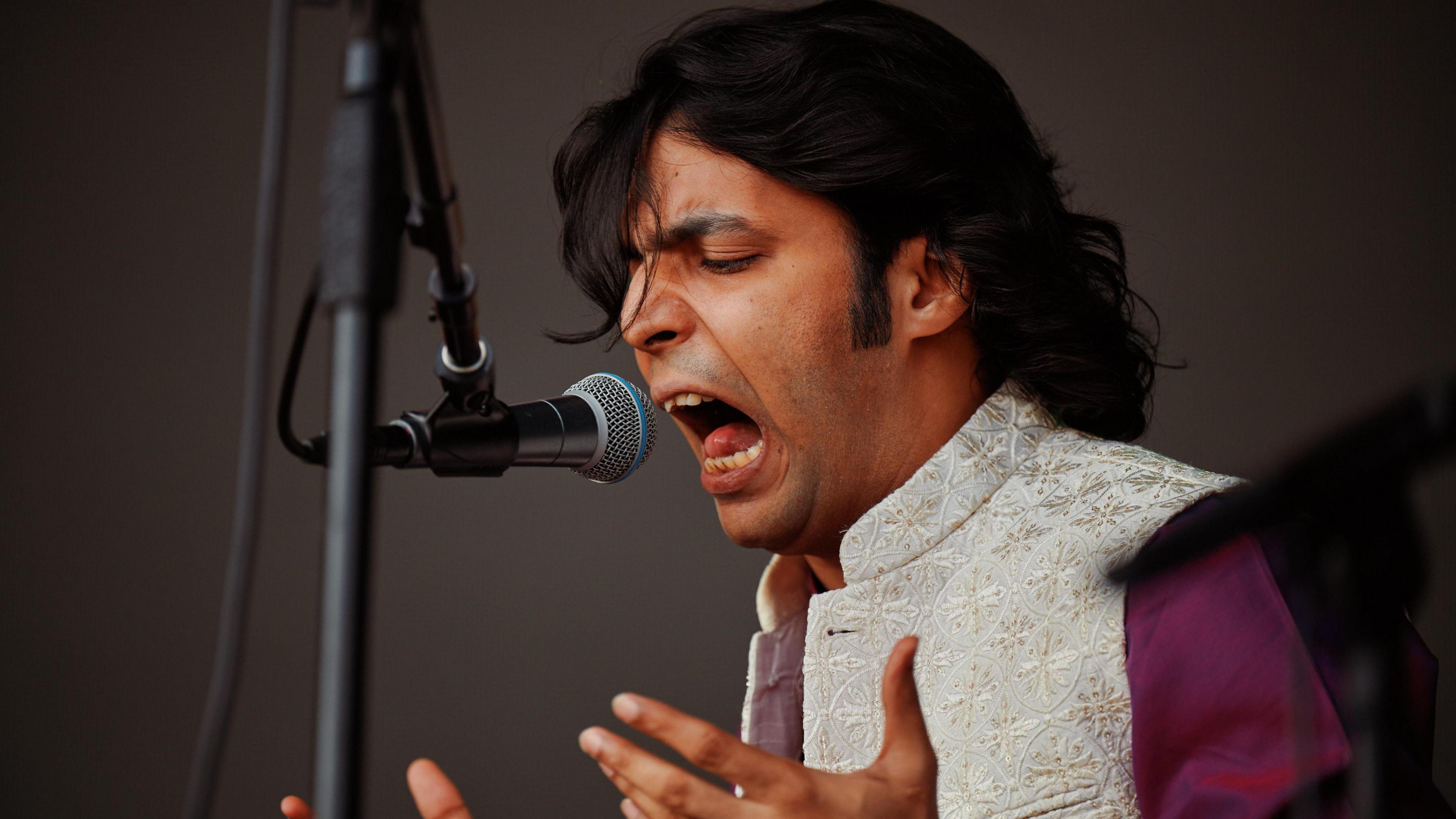 A person with dark hair wearing a ivory coloured embroidered tunic singing passionately into a microphone