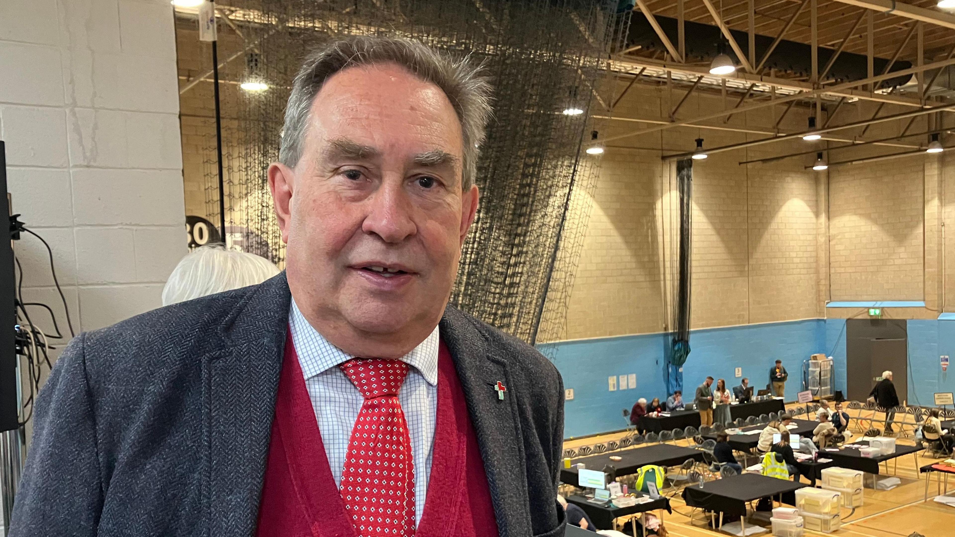 Stroud MP David Drew. He is wearing a grey suit jacket with a blue and white shirt, a red tie and red jumper underneath. He is stood on a balcony above a local election count.