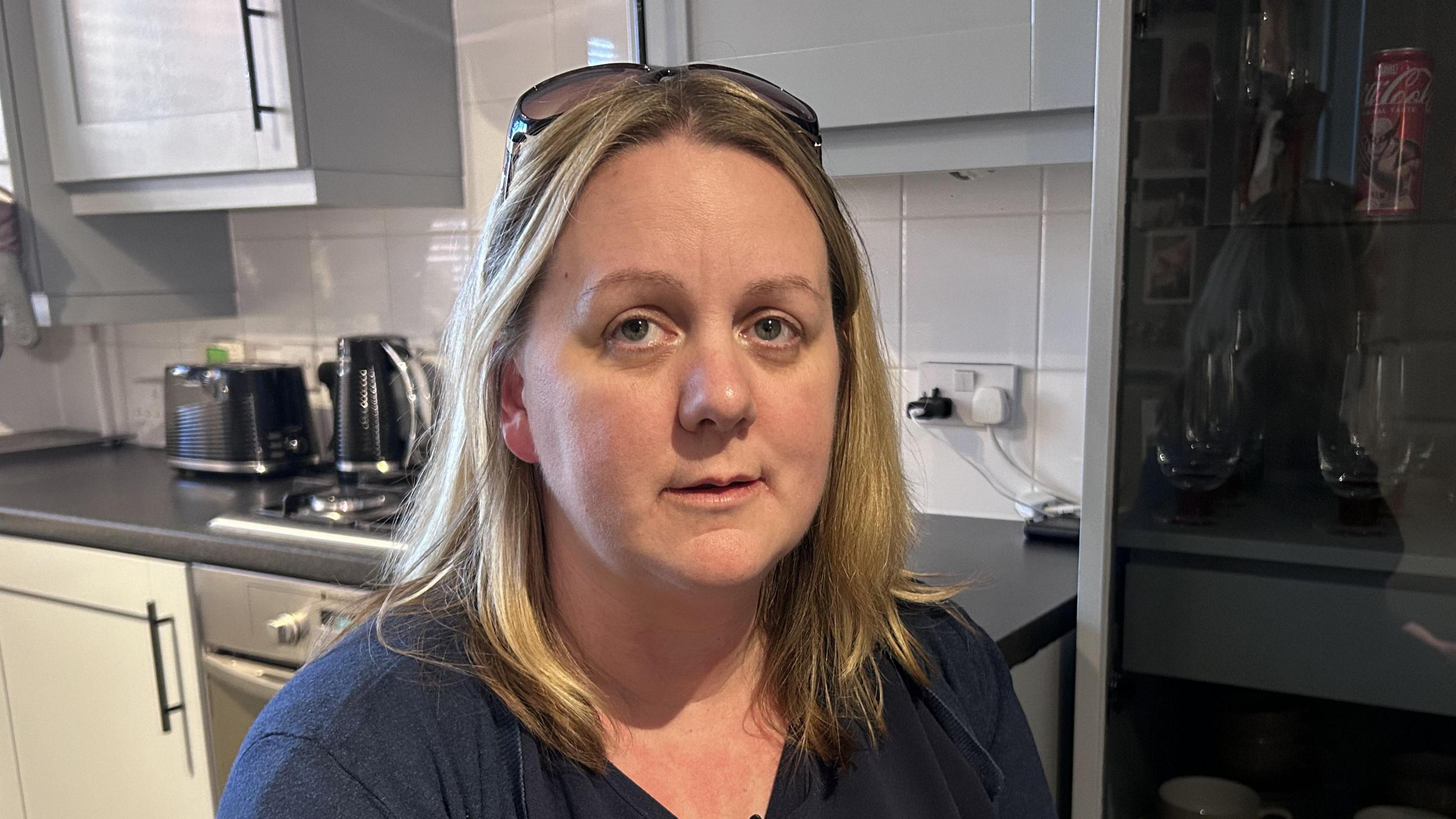 Rosemary with below shoulder length blonde hair sits in her kitchen, Behind her is a work surface. She has glasses on her head and wears a short sleeve navy blue t-shirt.