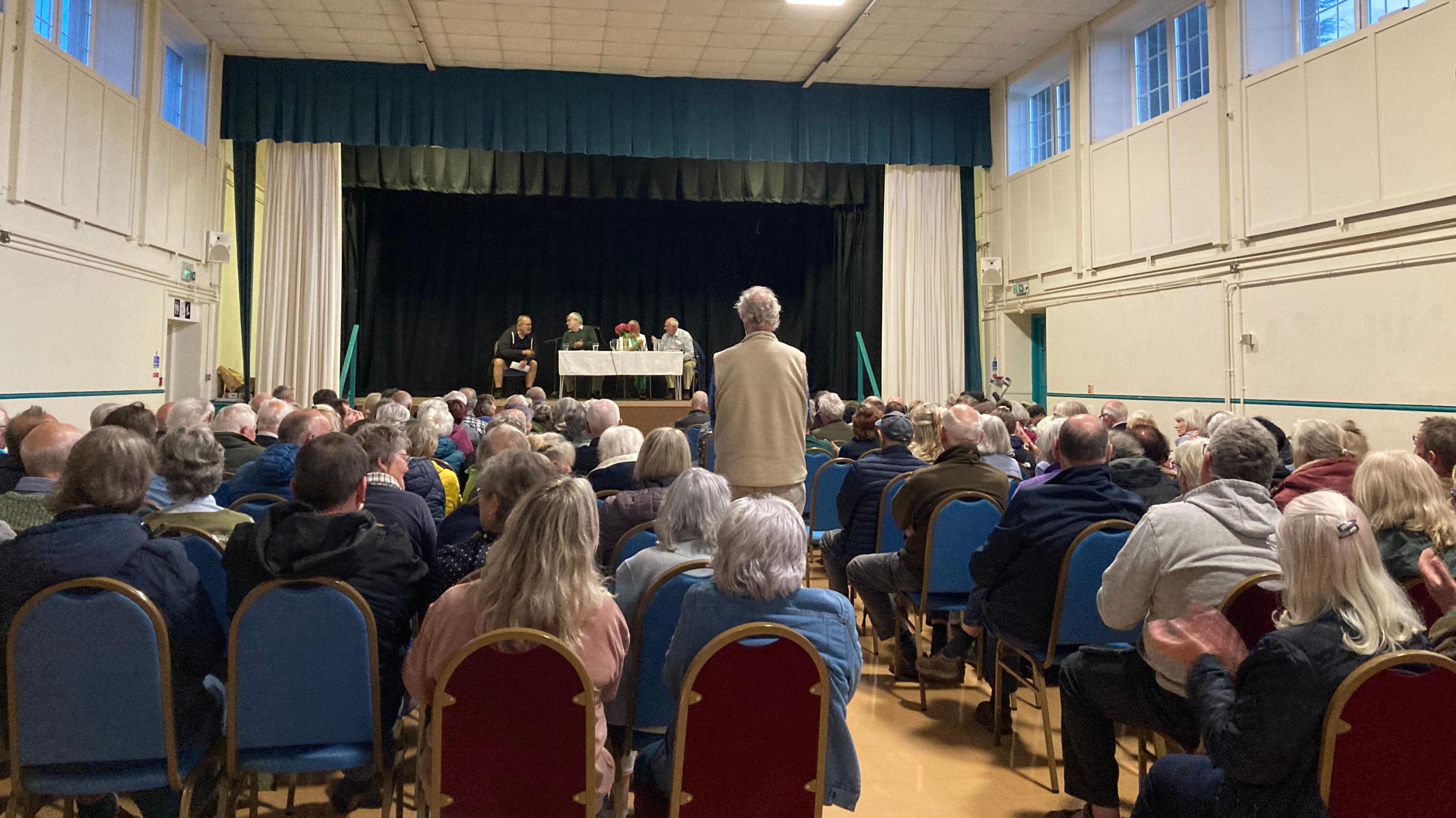 People sat in rows in a community hall with their backs to the camera, and facing a stage on which four people sit behind a table