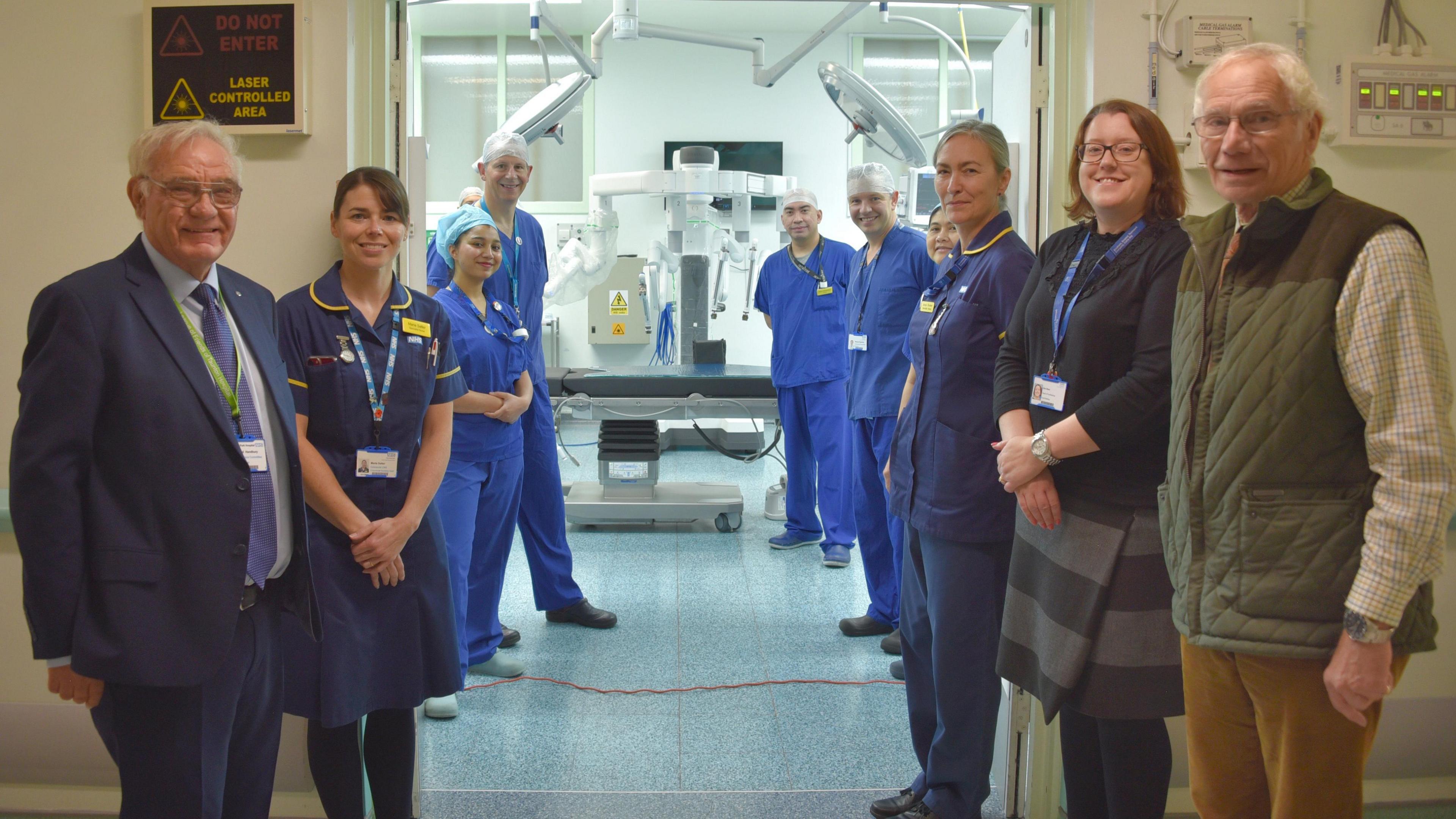 Staff members at Musgrove Park Hospital are standing next to each other in front of an operating room. Some are wearing blue scrubs. They are all looking at the camera and smiling.