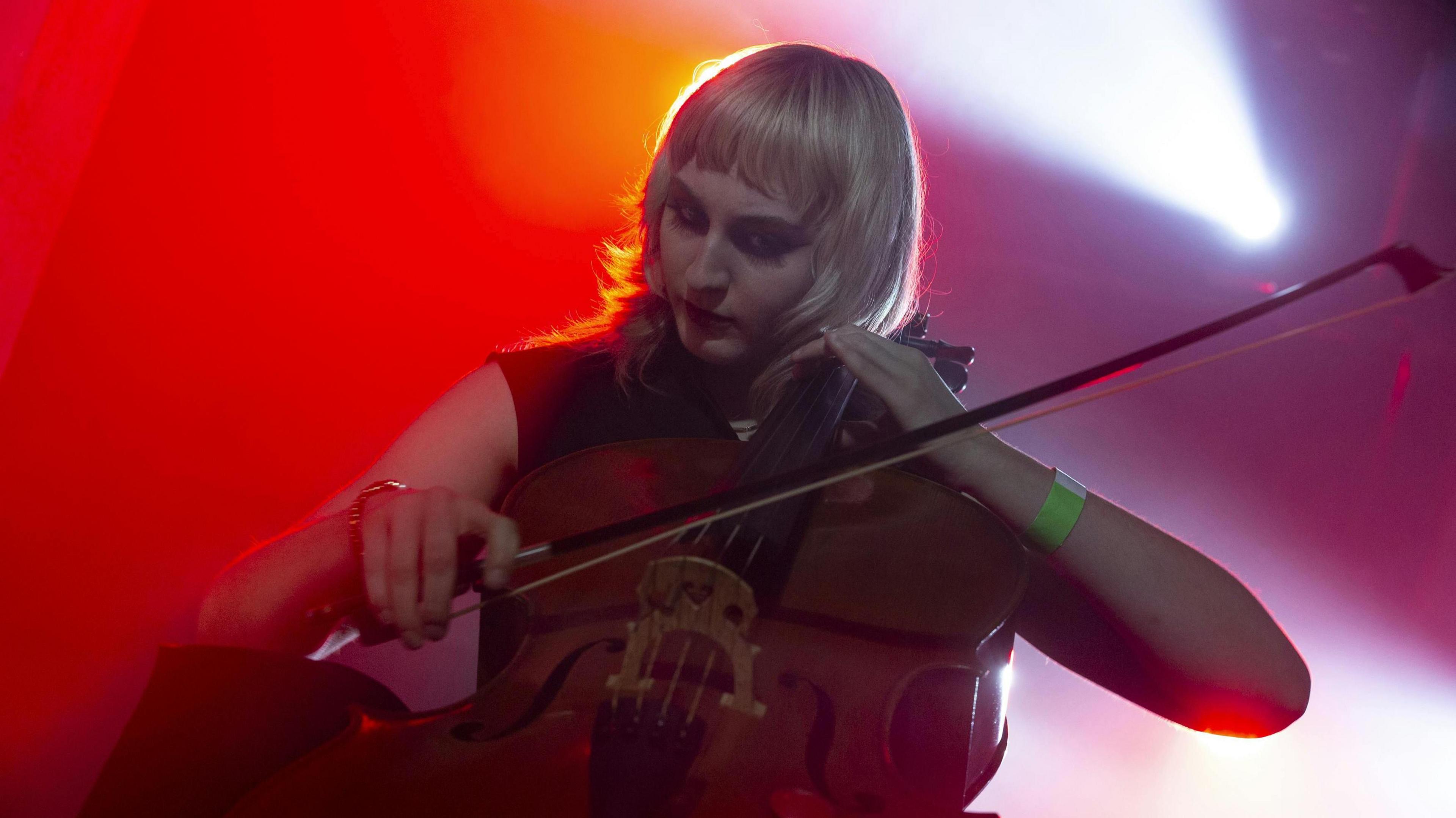 Olivia McLean playing cello onstage at a Tenementals gig. She has blonde hair, while the stage is bathed in red light