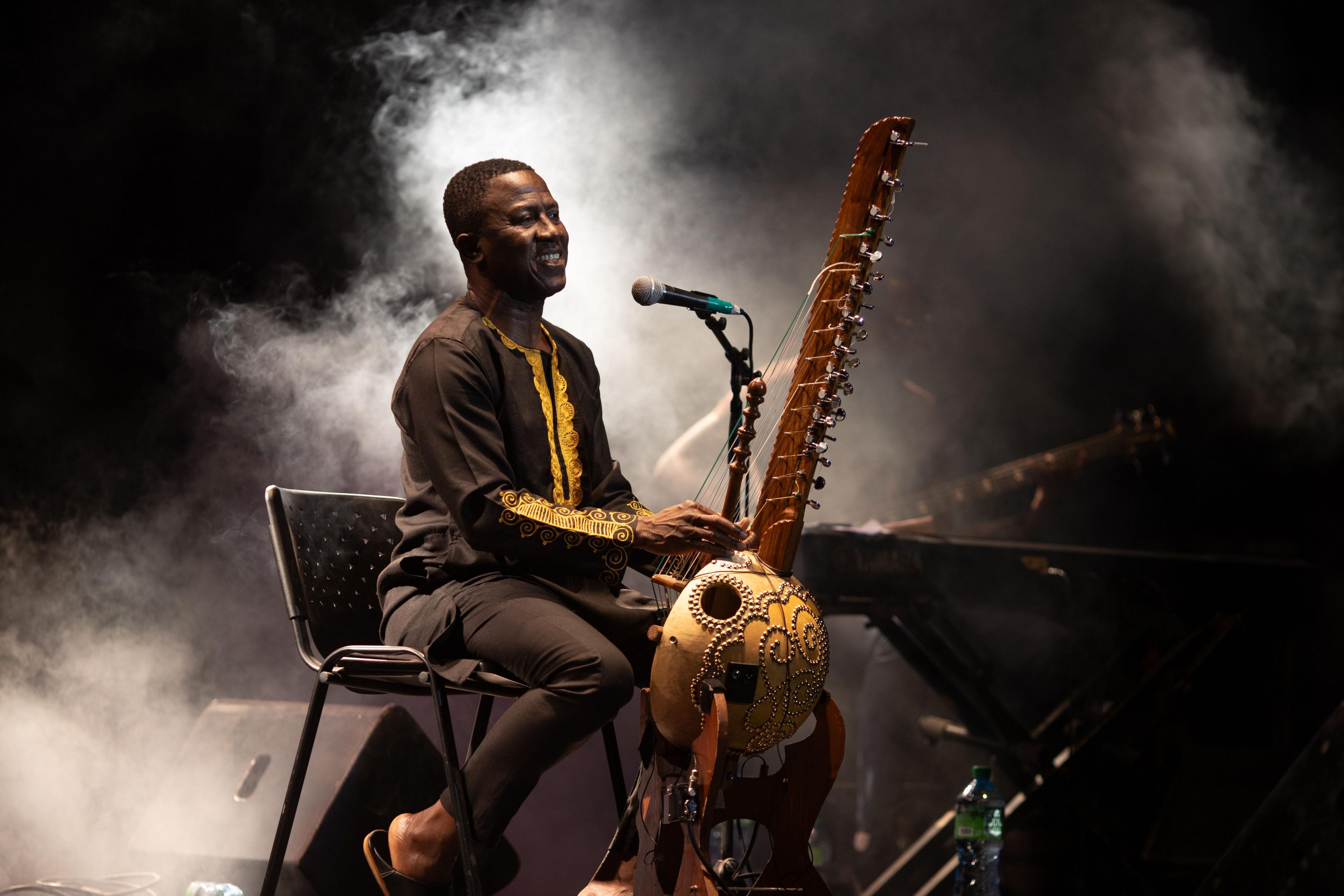 Smoke is in the background as Lamine Cissoko smiles whilst he plays the kora.