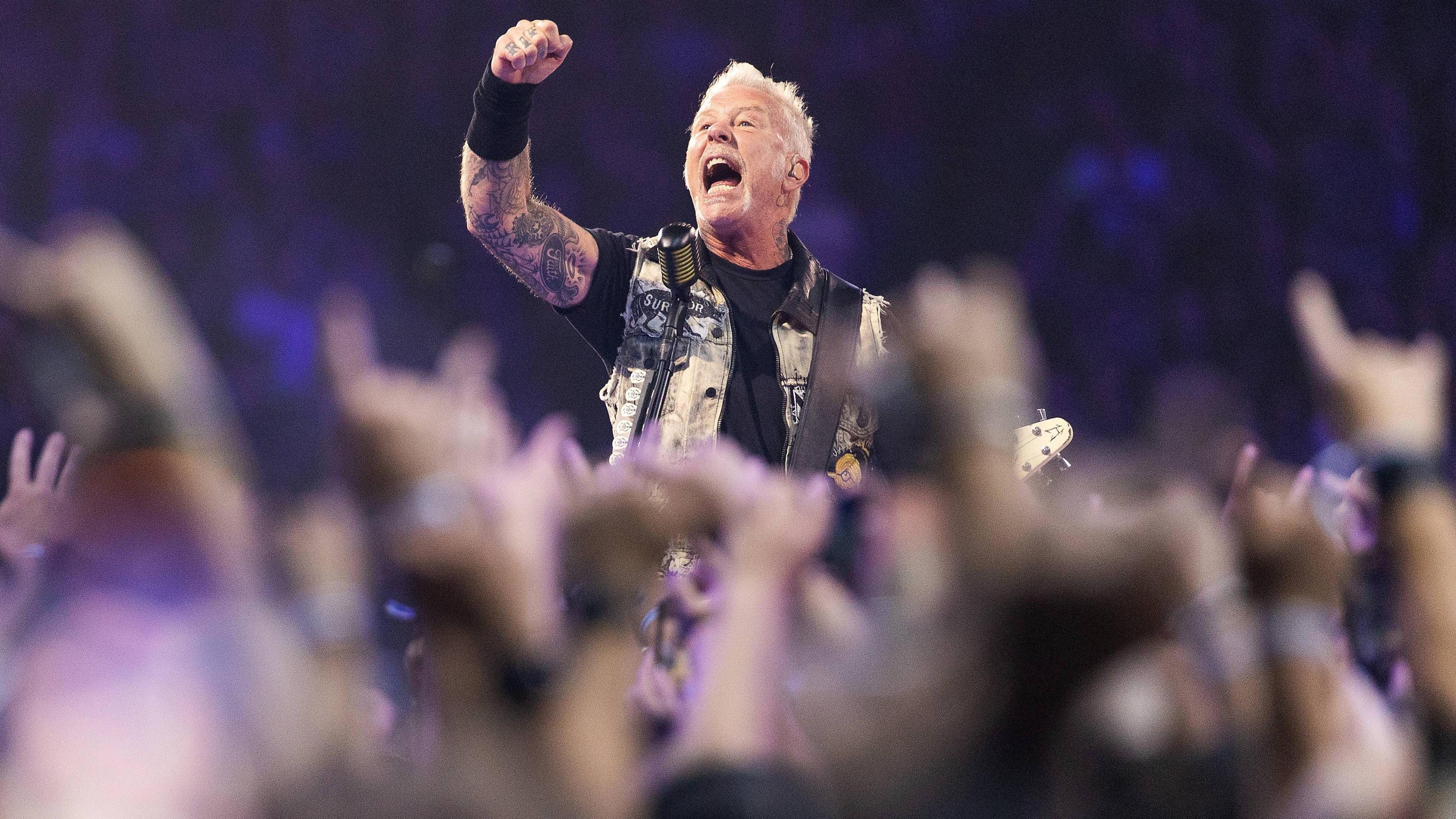 Lead singer and guitarist of Metallica, James Hetfield performs onstage at Lumen Field. He's surrounded by a crowd of people with their arms up. He's wearing a black t shirt, arm cuff and a denim vest. He's holding a guitar. He's got his arm up in the air and appears to be shouting.