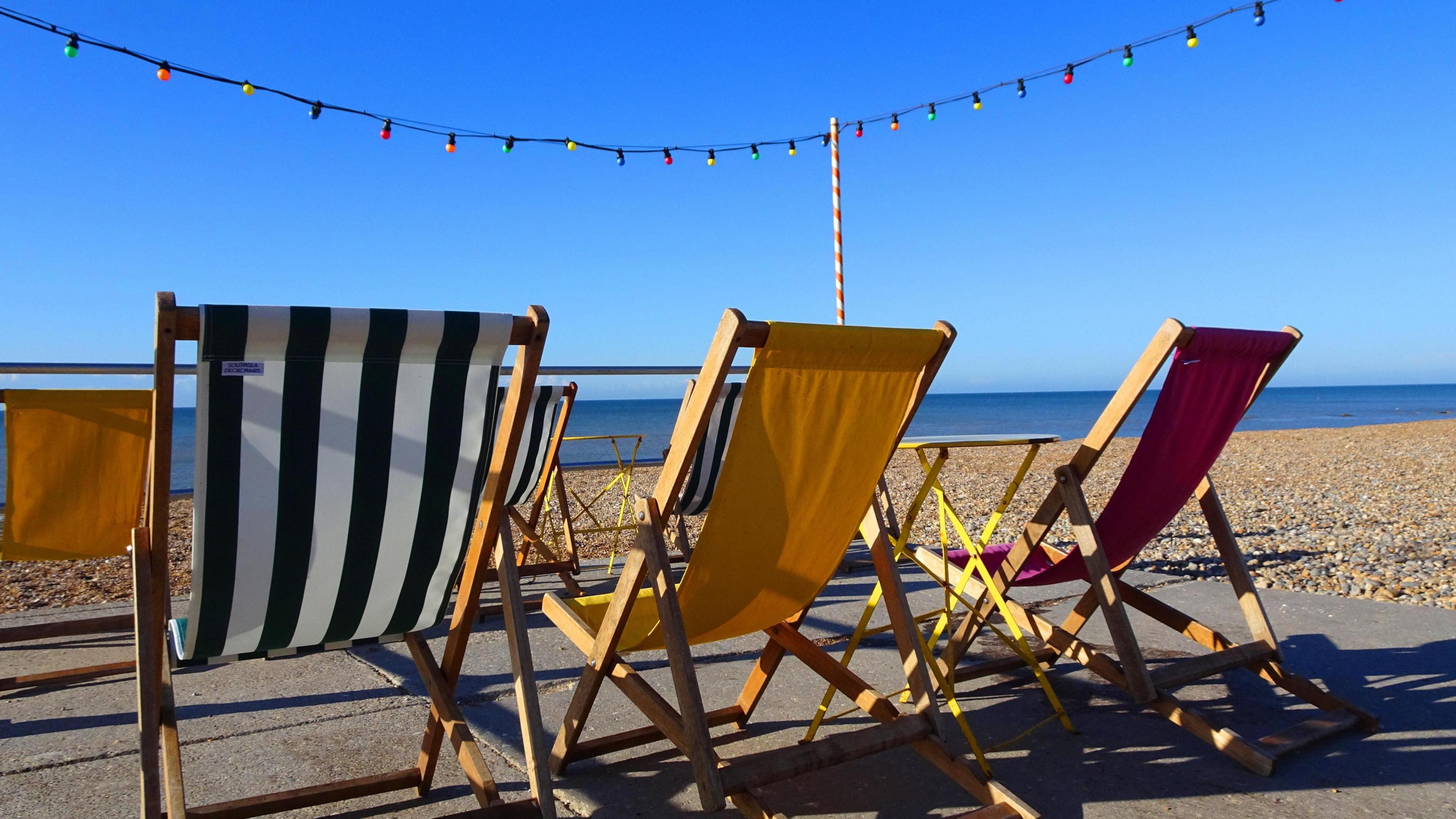 Picture of a clear sunny day on the beach with three unoccupied deckchairs.