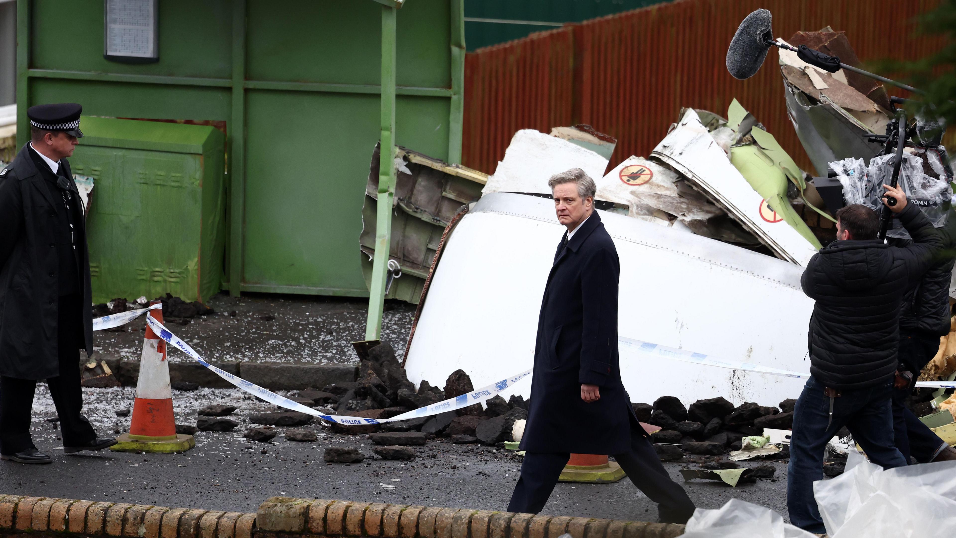 Colin Firth plays Dr Jim Swire in a new TV series. The photo shows Colin Firth in character, in a black suit and tie, walking down a path. Behind him is plane rubble and police tape. A man dressed in a police uniform stands to the left-hand side of the image, and a man operating a camera and a mic stands behind Firth.