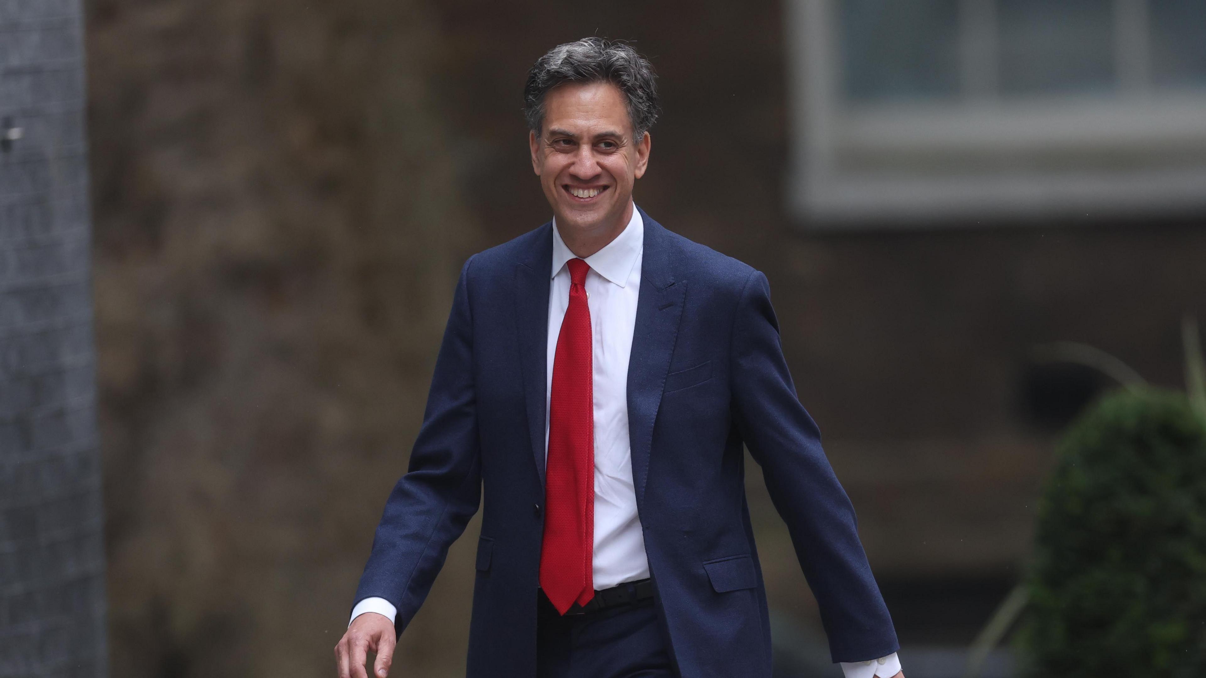 A smiling Ed Miliband wearing a navy blue suit with a red tie is pictured walking down on Street