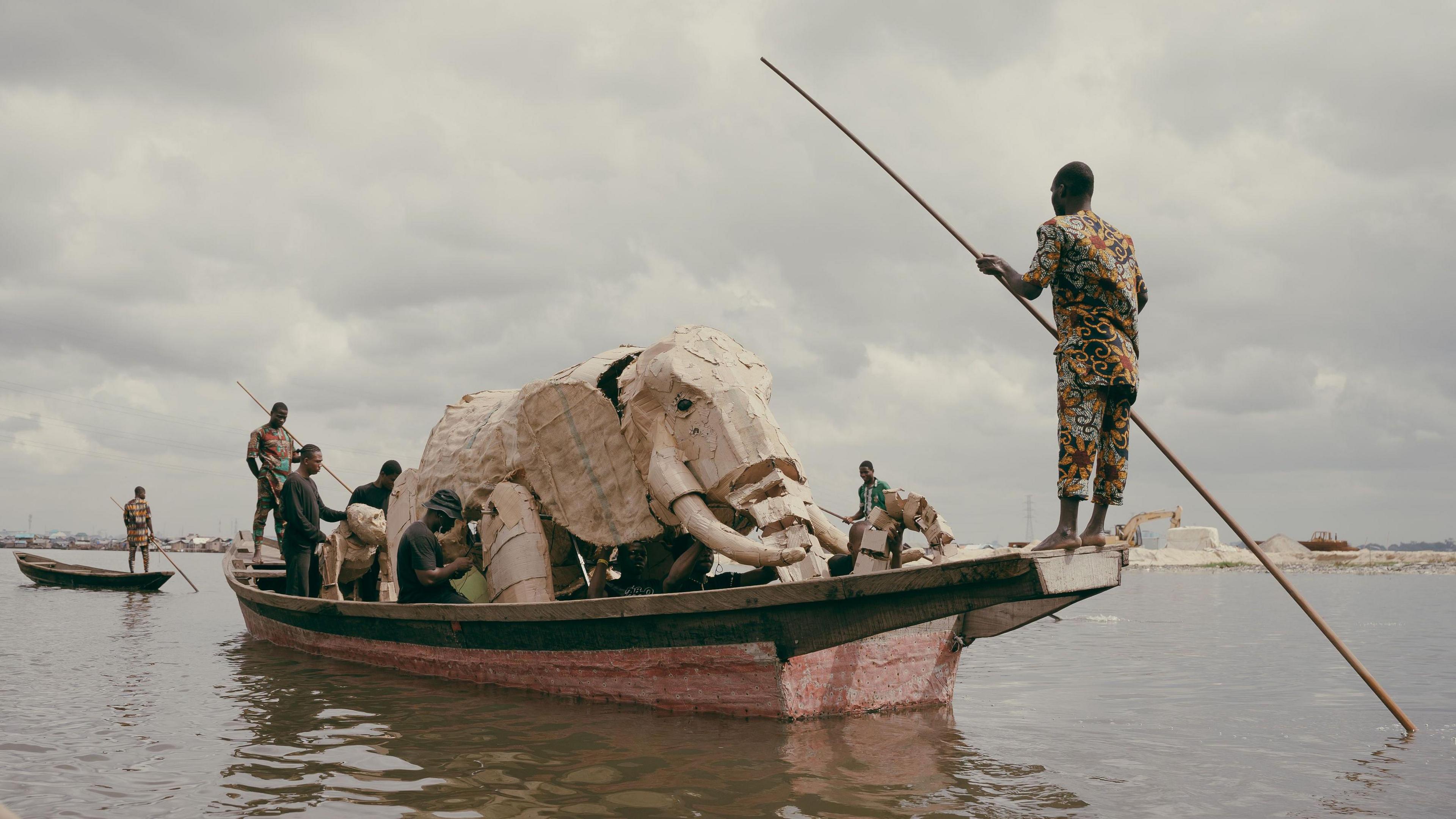A large puppet elephant and ape being transported on a small boat on the Makoko River in Lagos. There are a number of people on the boat supporting the animals. One man is stood at the front of the boat with a large stick in the water.