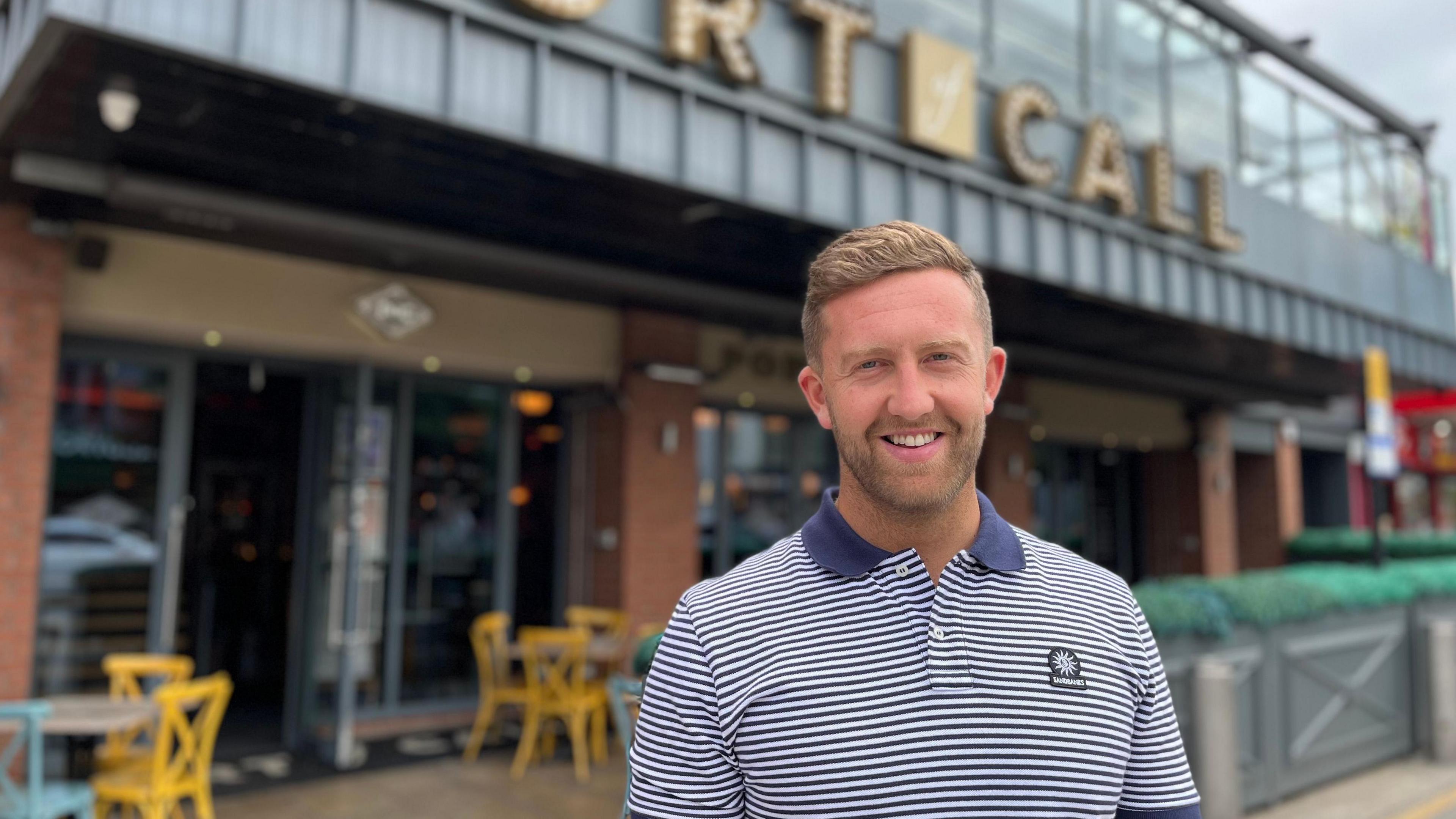 Adam Dickman stands in front of a pub he owns in Sunderland city centre called Port of Call. He is wearing a navy blue and white striped polo shirt. 