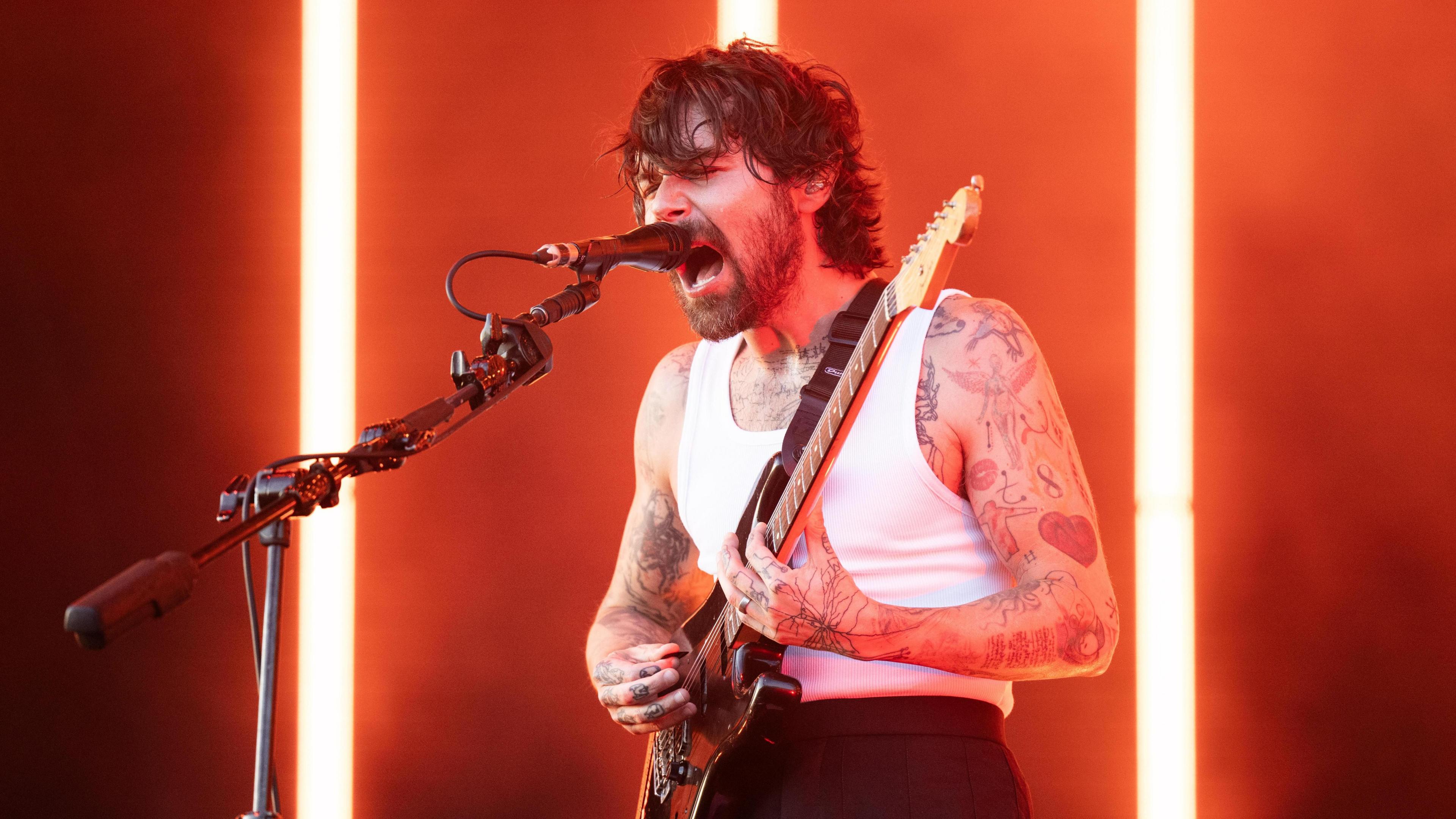 Simon Neil of Biffy Clyro onstage -he is wearing a vest while playing the guitar and singing into a microphone.
He is bearded and has tattoos over his arms.