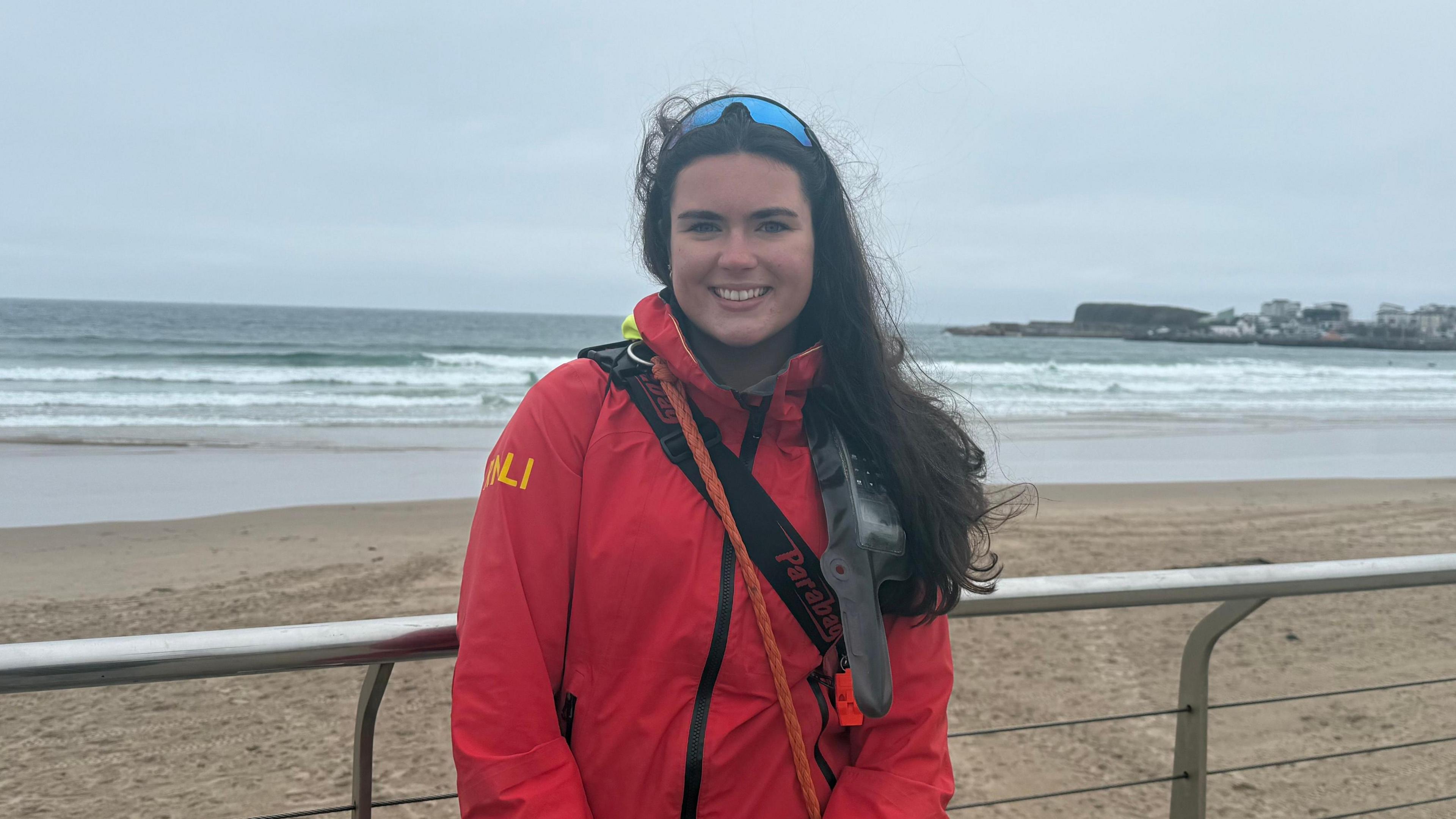 Maria Dillon. She has long dark hair wearing a red RNLI lifeguard uniform and sunglasses on her head. She is standing by railings with the see in the background.