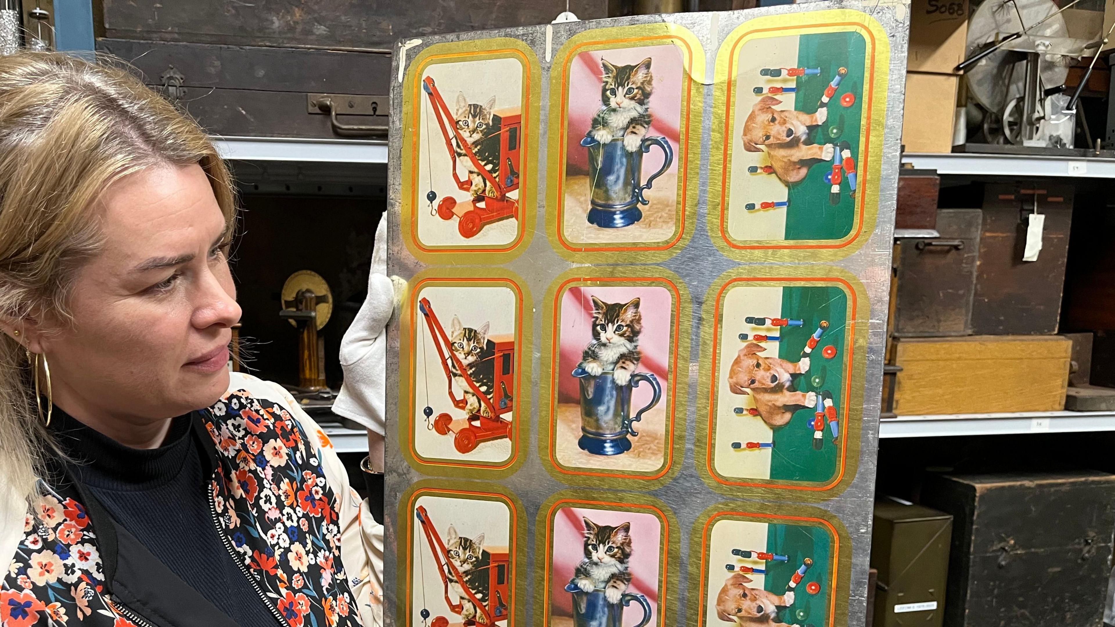 A woman with blonde hair holds a metal sheet with pictures of kittens in jugs, kittens on toy cranes and a puppy playing with toy soldiers.